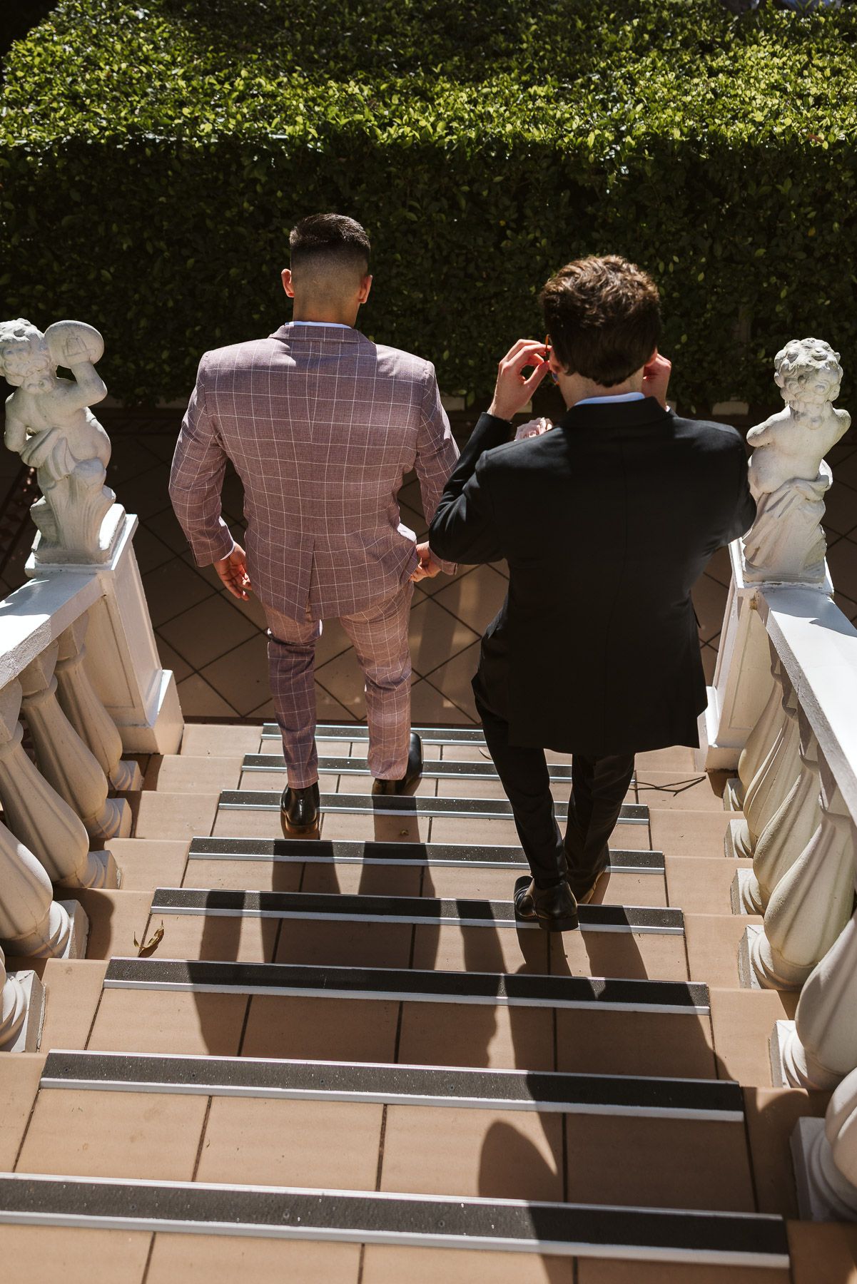 Two men in suits walking up elegant stone stairs with classical statues and green hedges in sunlight.