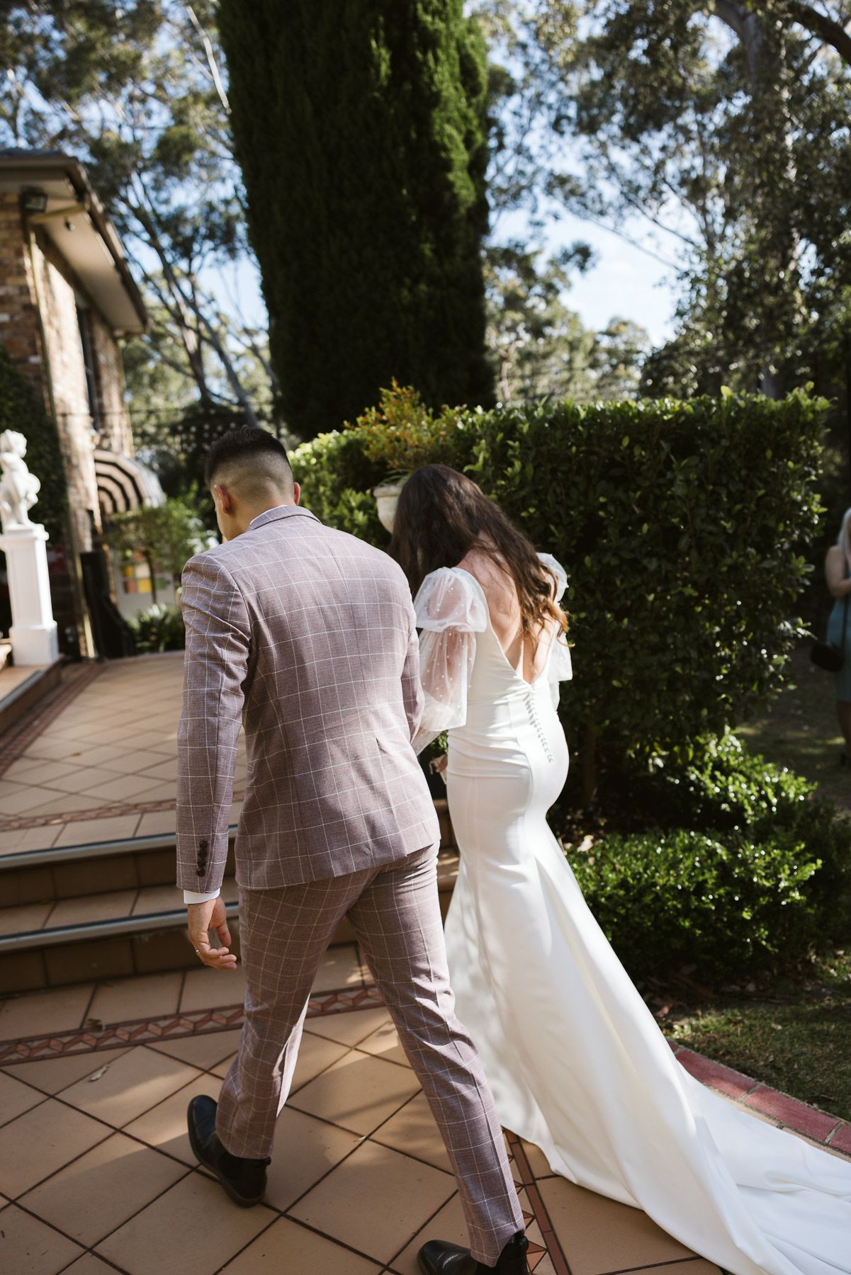 A bride in a white gown and groom in a plaid suit walk hand in hand through a lush garden venue.