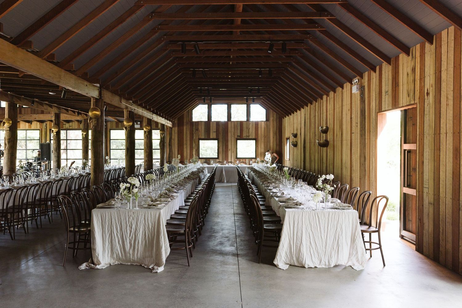 Rustic barn wedding venue with long banquet tables set up under wooden beams and natural lighting.