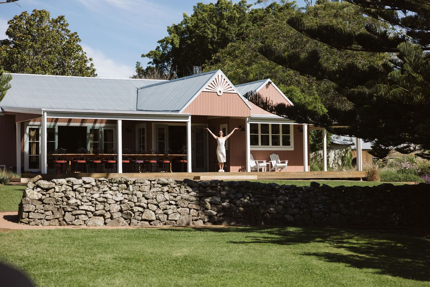 Modern farmhouse with wrap-around porch and stone retaining wall set against lush green landscape.