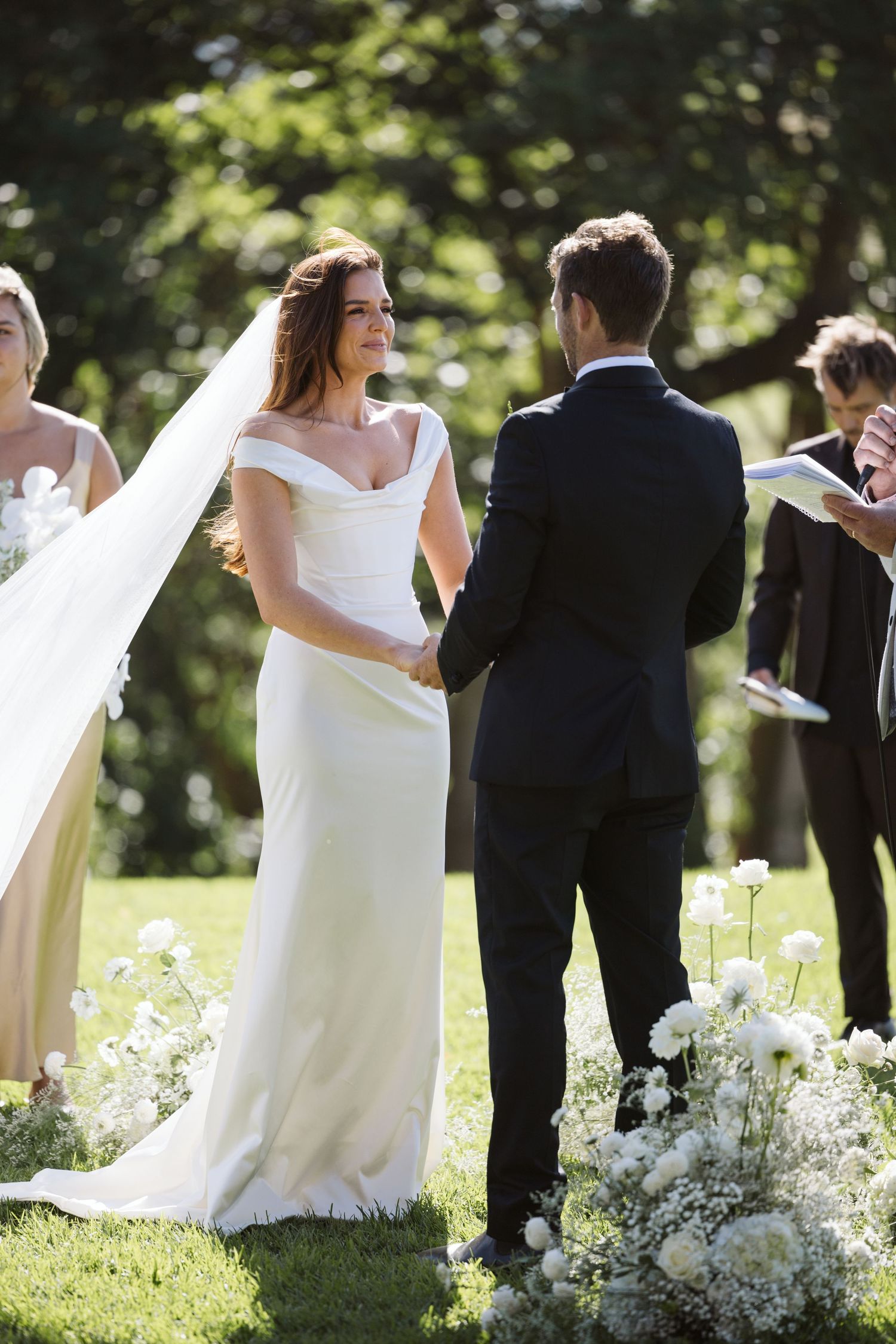 Outdoor wedding ceremony amidst trees with white floral arrangements on the ground.