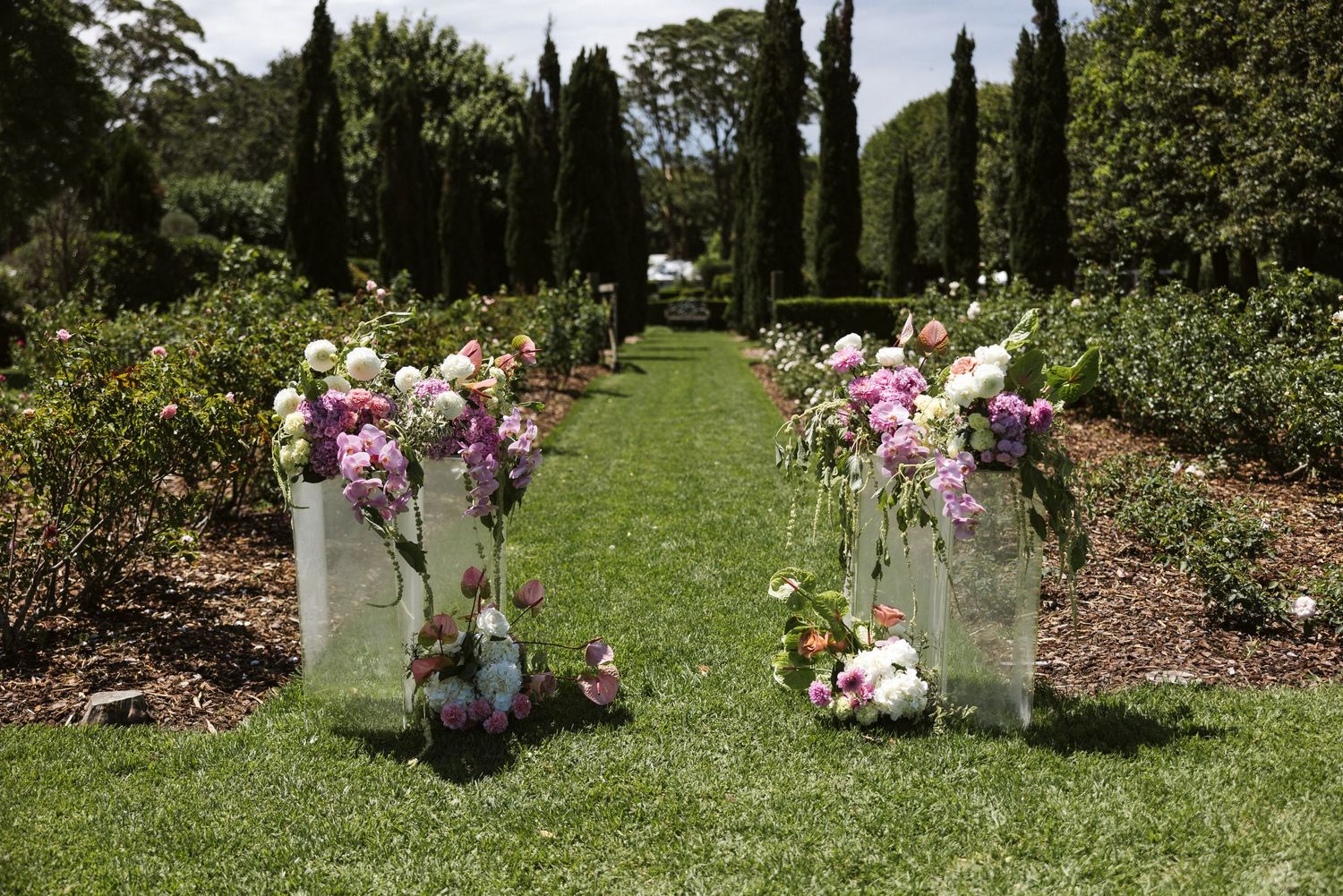 Garden ceremony setup with glass vases filled with pink and purple flowers lining a grass aisle.