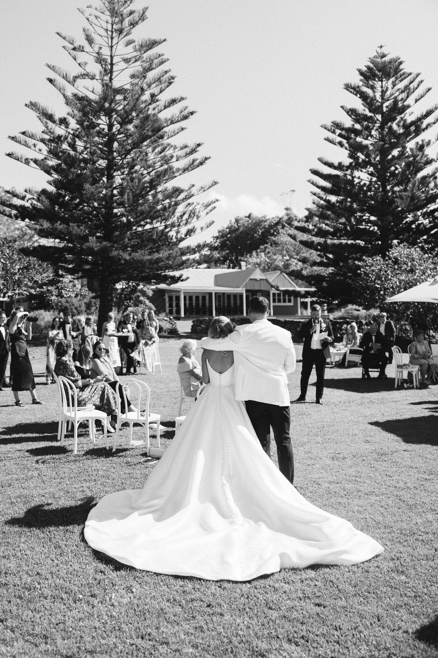 Black and white photo of newlyweds dancing outdoors with tall pine trees and guests in background.