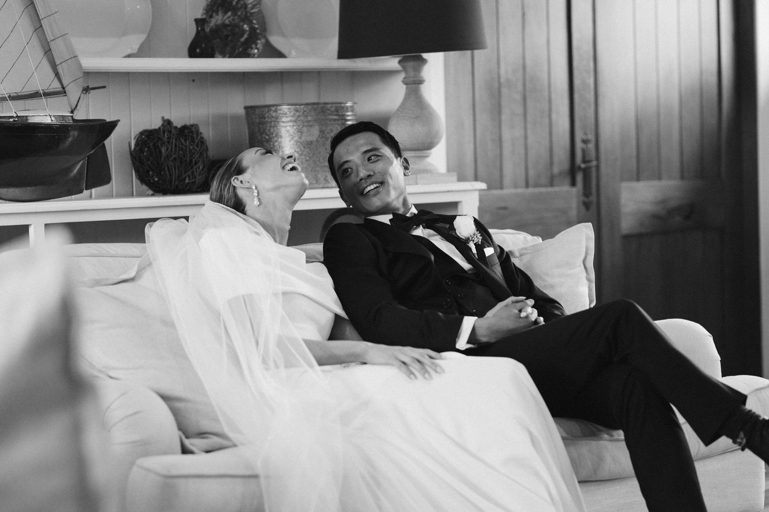Black and white candid photo of newlyweds relaxing on white couch in formal attire.