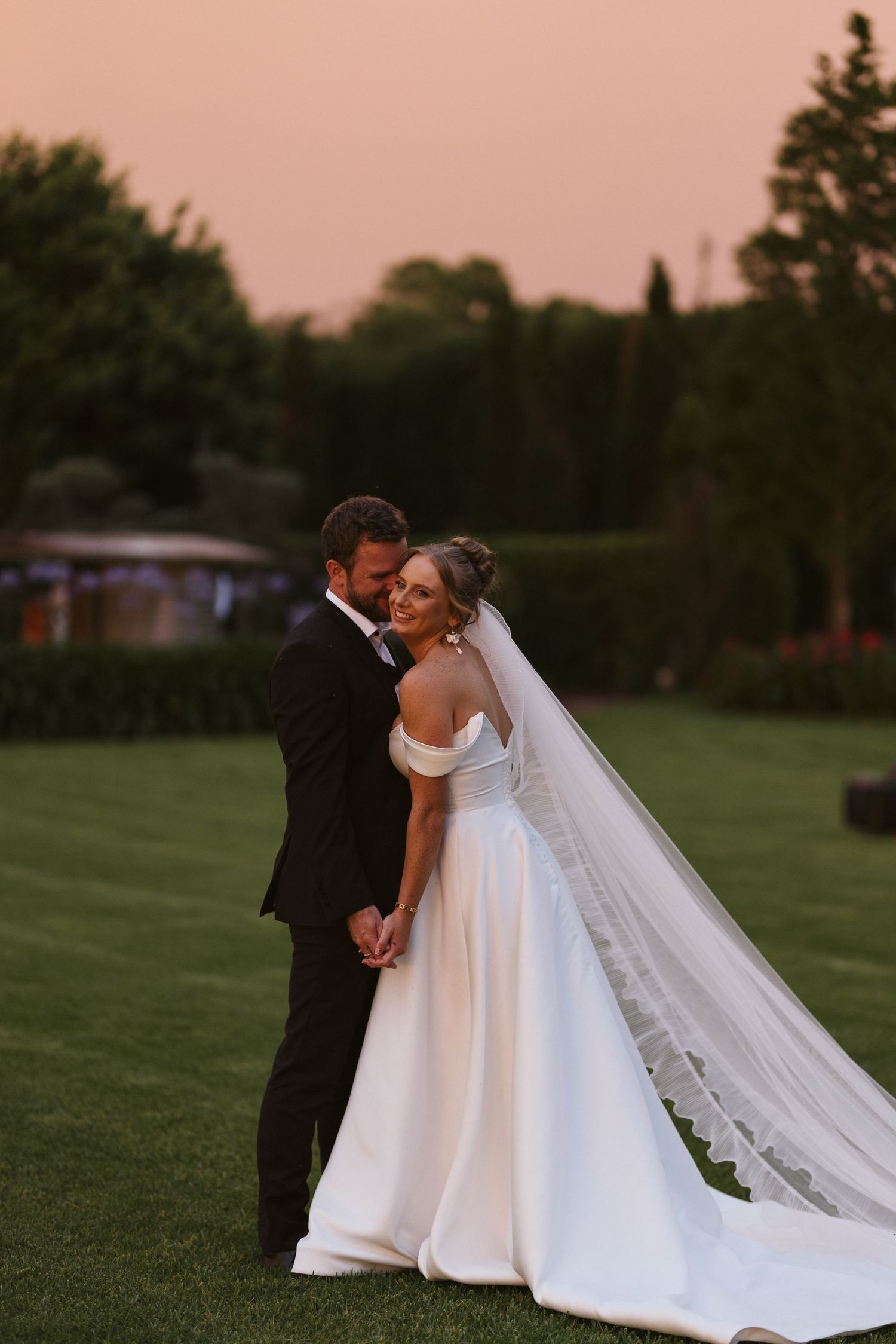 Sunset wedding portrait with couple embracing on green lawn with warm pink sky.