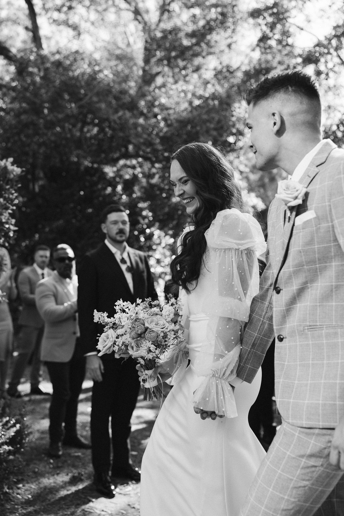 Black and white photo of smiling bride holding bouquet walking with groom at outdoor wedding ceremony.
