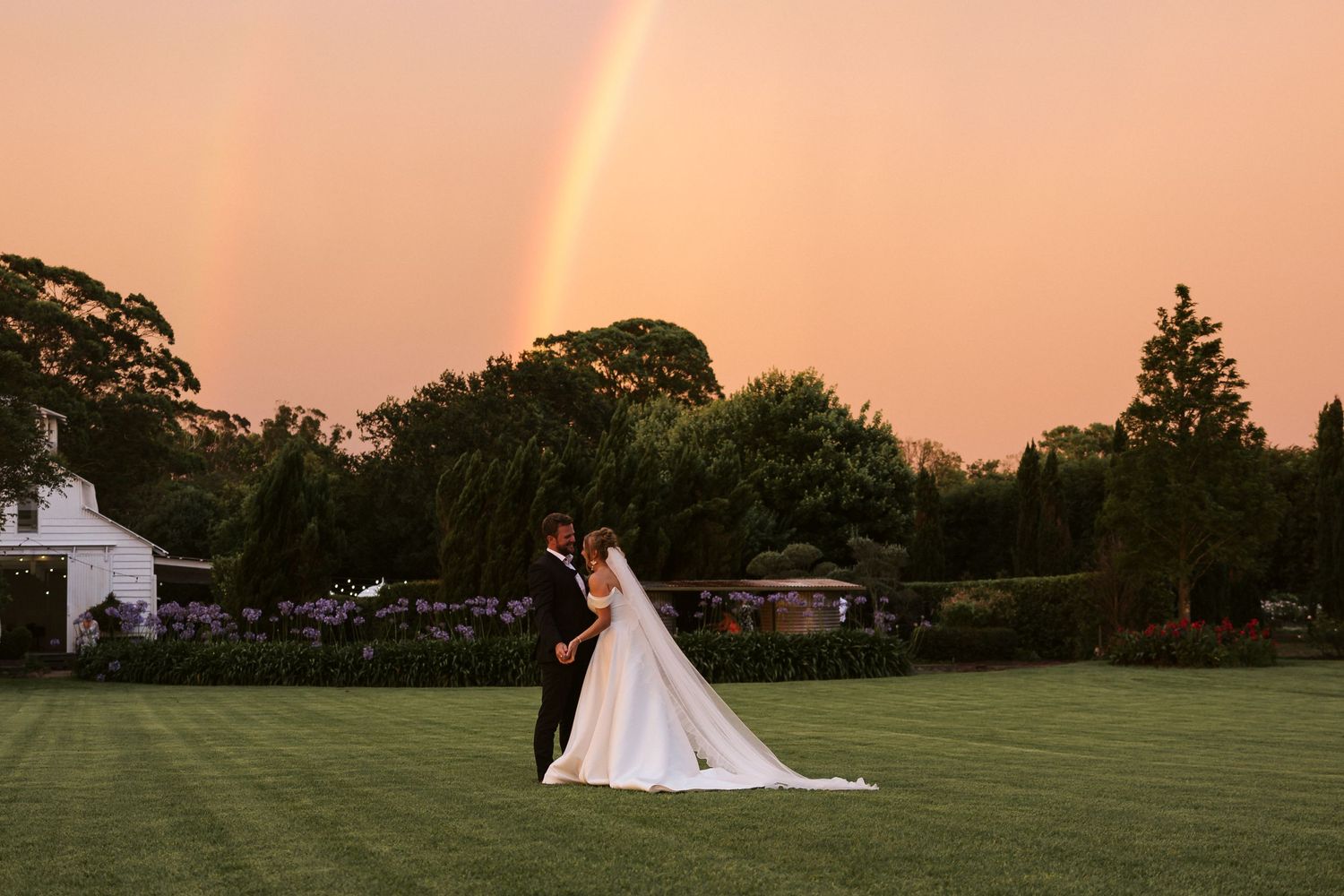 Wedding couple in garden with double rainbow in sunset sky.