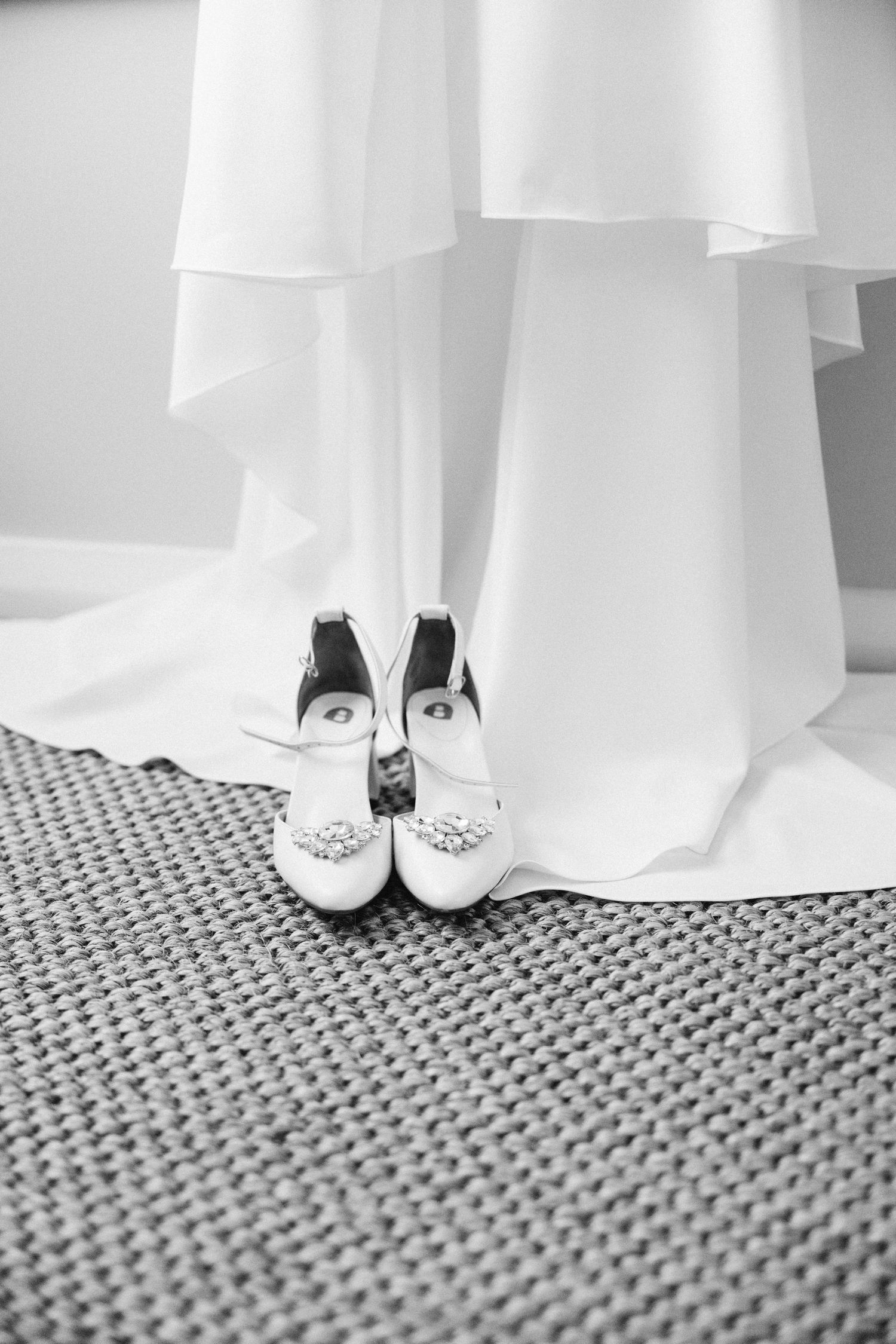 Elegant white bridal shoes with crystal embellishments resting on a textured carpet beneath a flowing white dress.