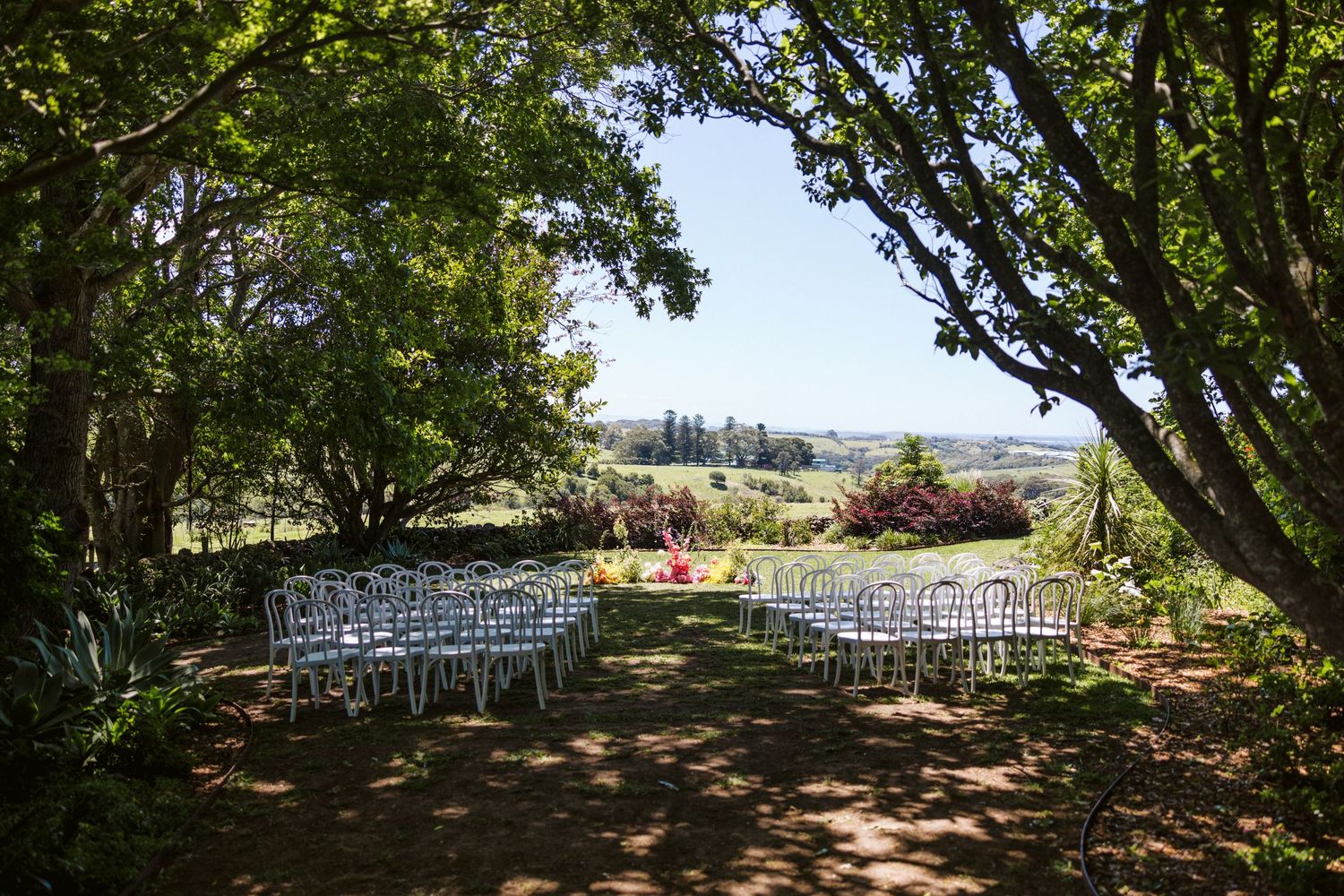 Outdoor wedding ceremony setup with white chairs framed by natural greenery.
