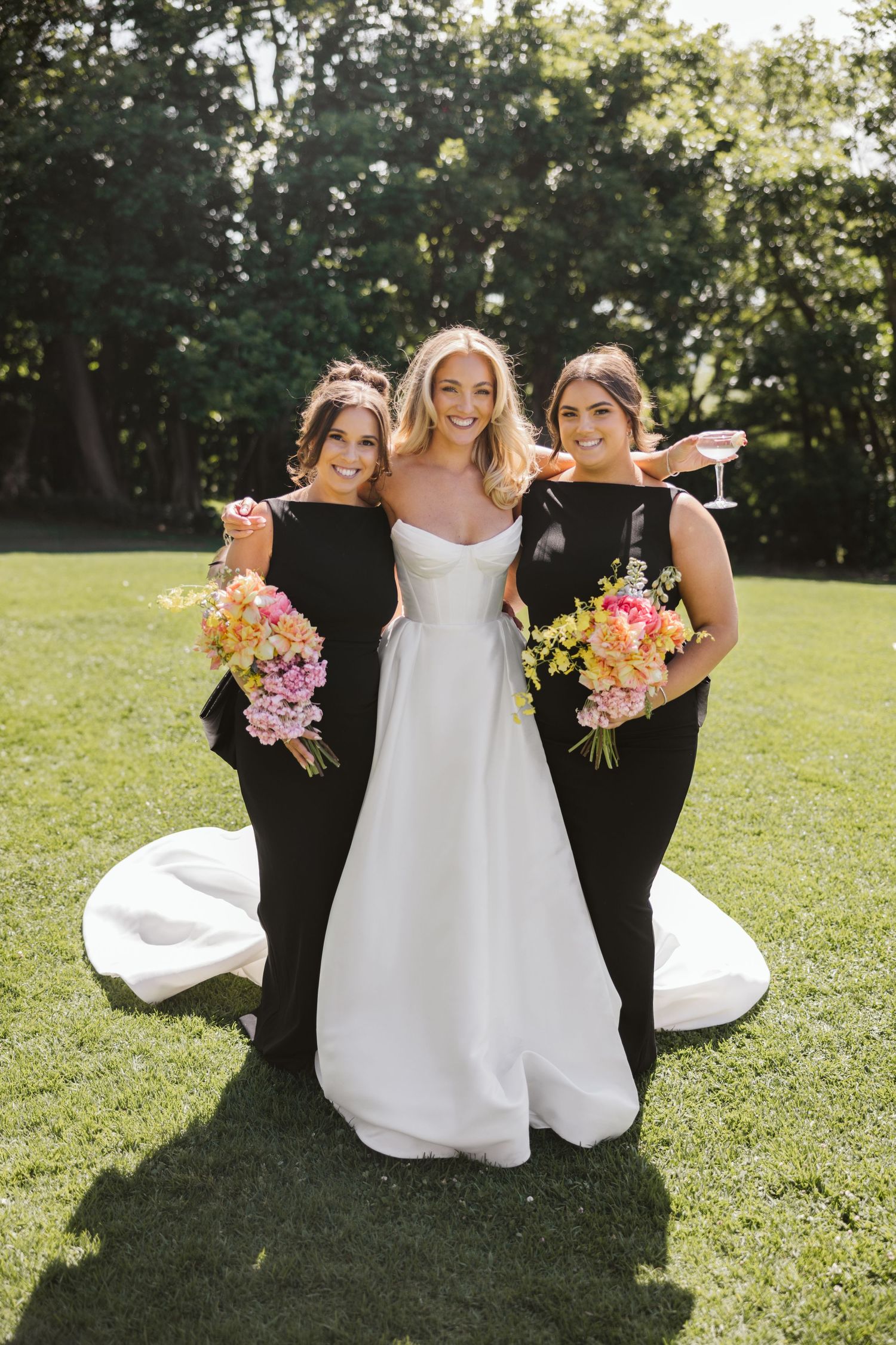 Bride in white gown poses with two bridesmaids in black dresses holding vibrant floral bouquets.