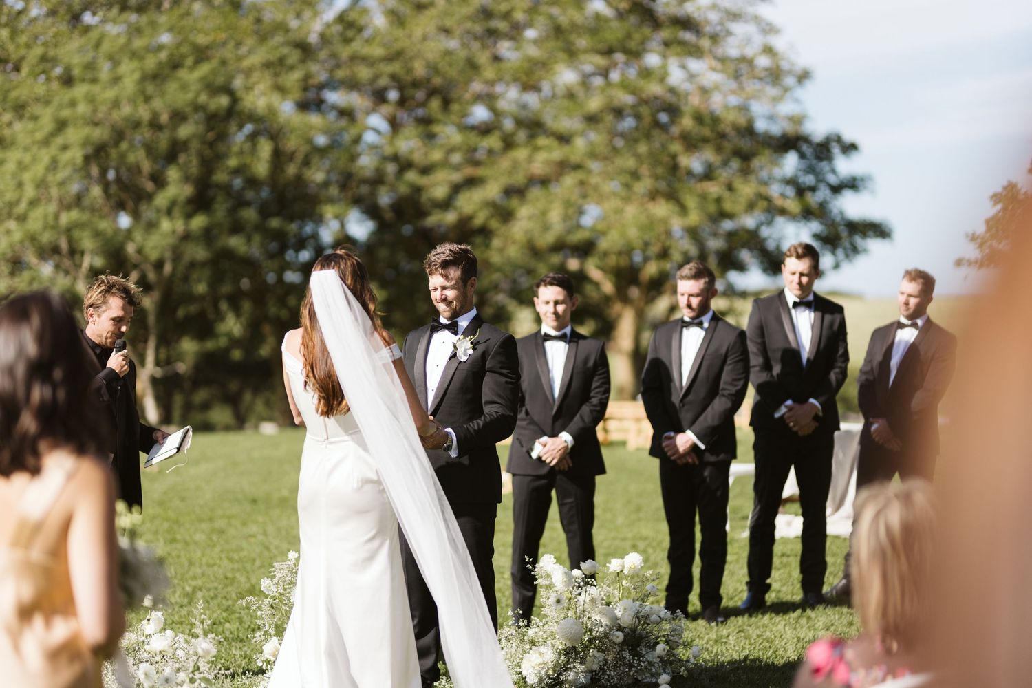 Wedding party stands in a line outdoors with groomsmen in dark suits.