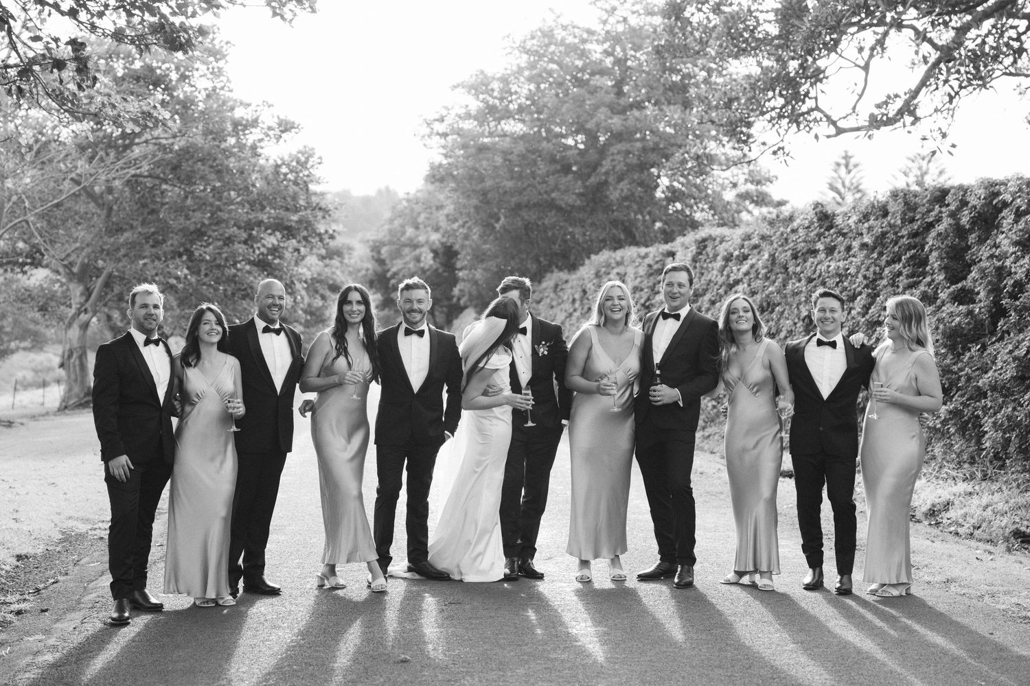 Black and white photo of wedding party lined up on tree-lined path.