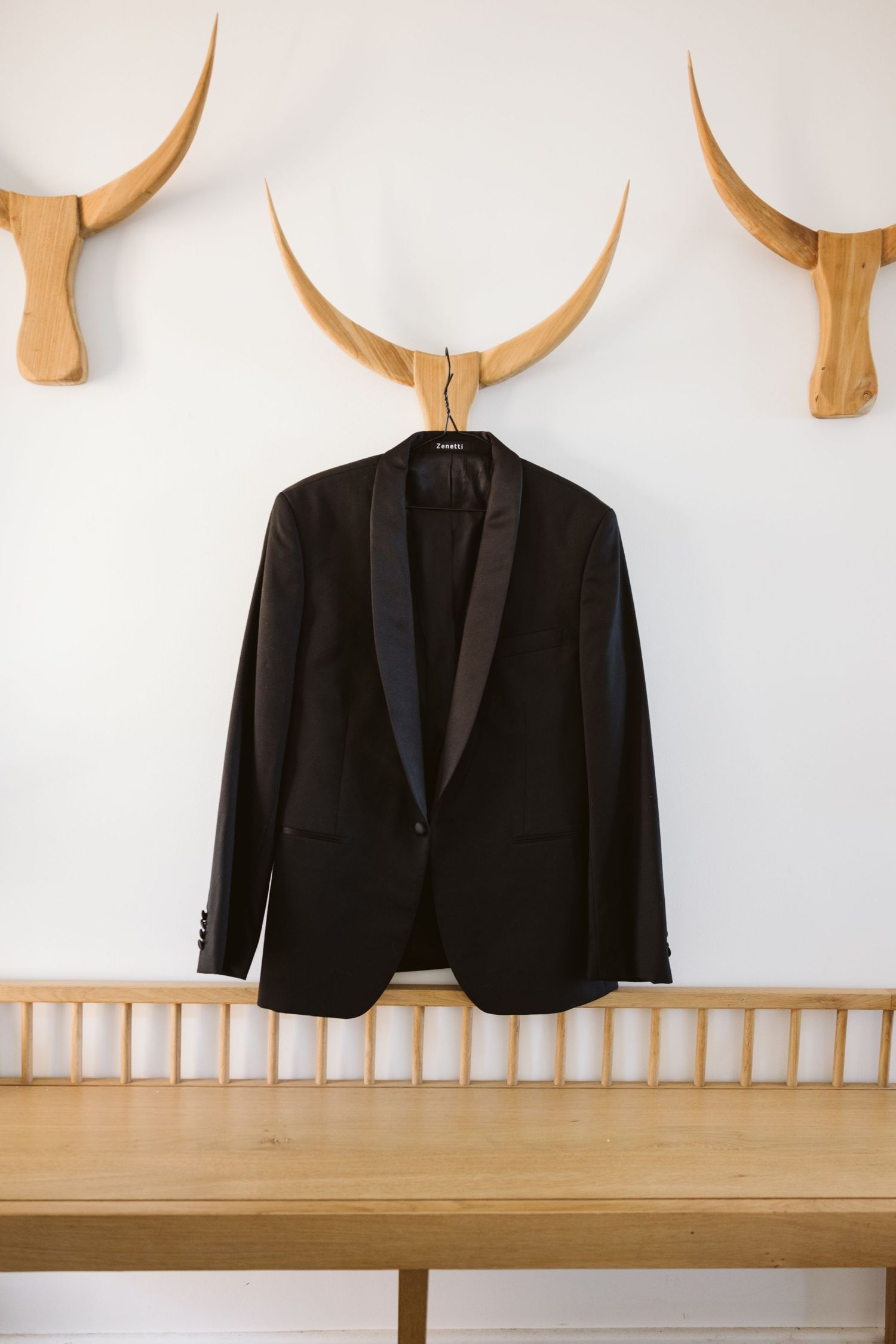 Black tuxedo jacket hanging on wooden bull horn wall mount above wooden bench.