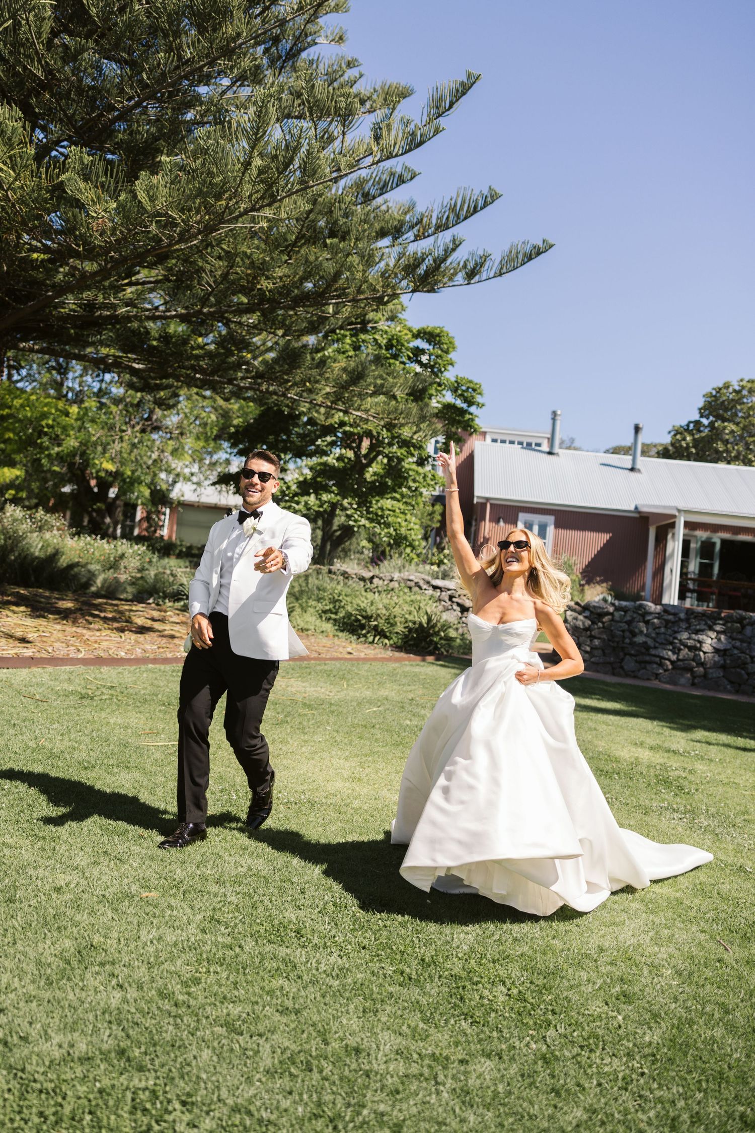Joyful couple dancing on green lawn with rustic building and pine trees in background.