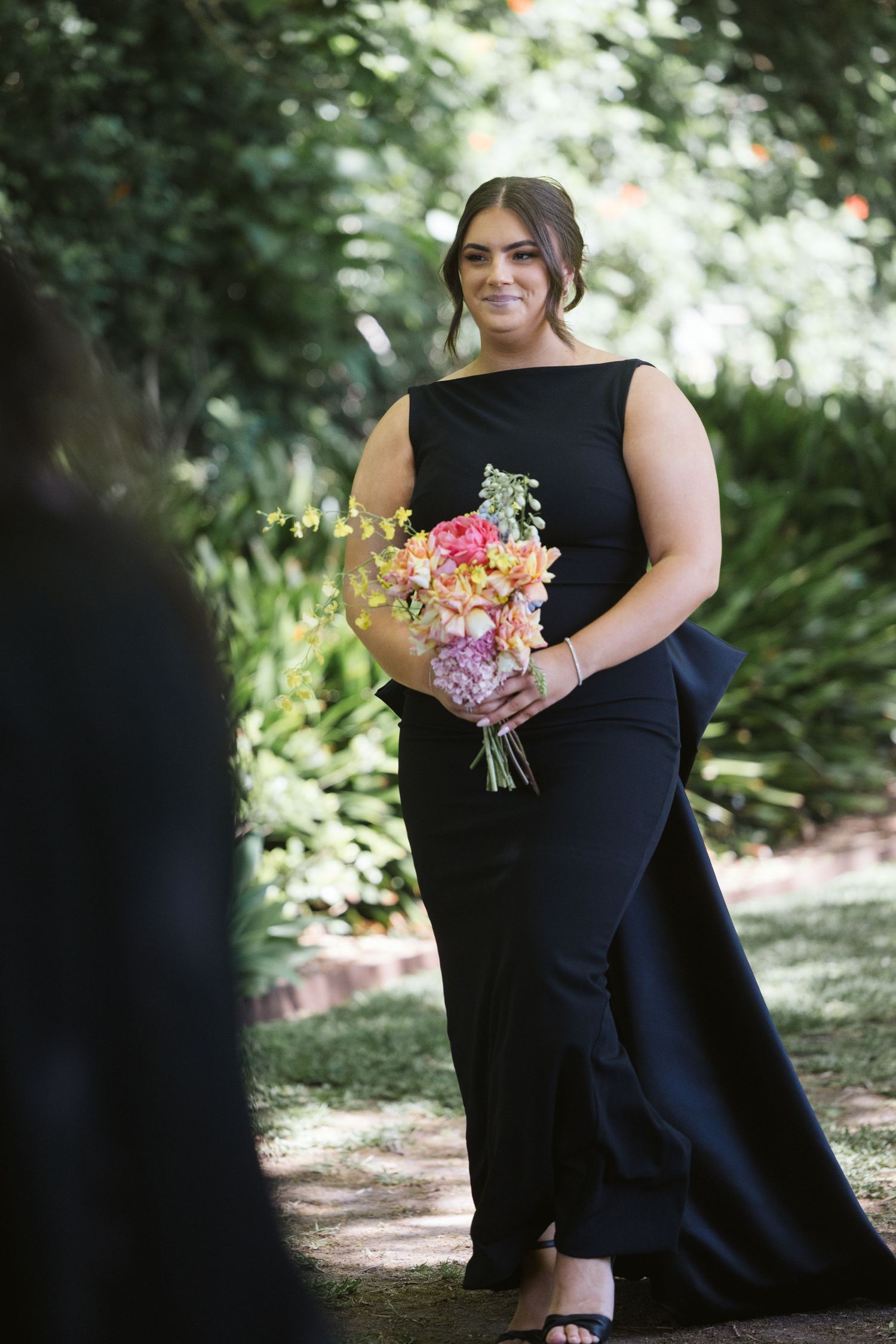 Bridesmaid in elegant black dress holds colorful spring bouquet in outdoor garden setting.