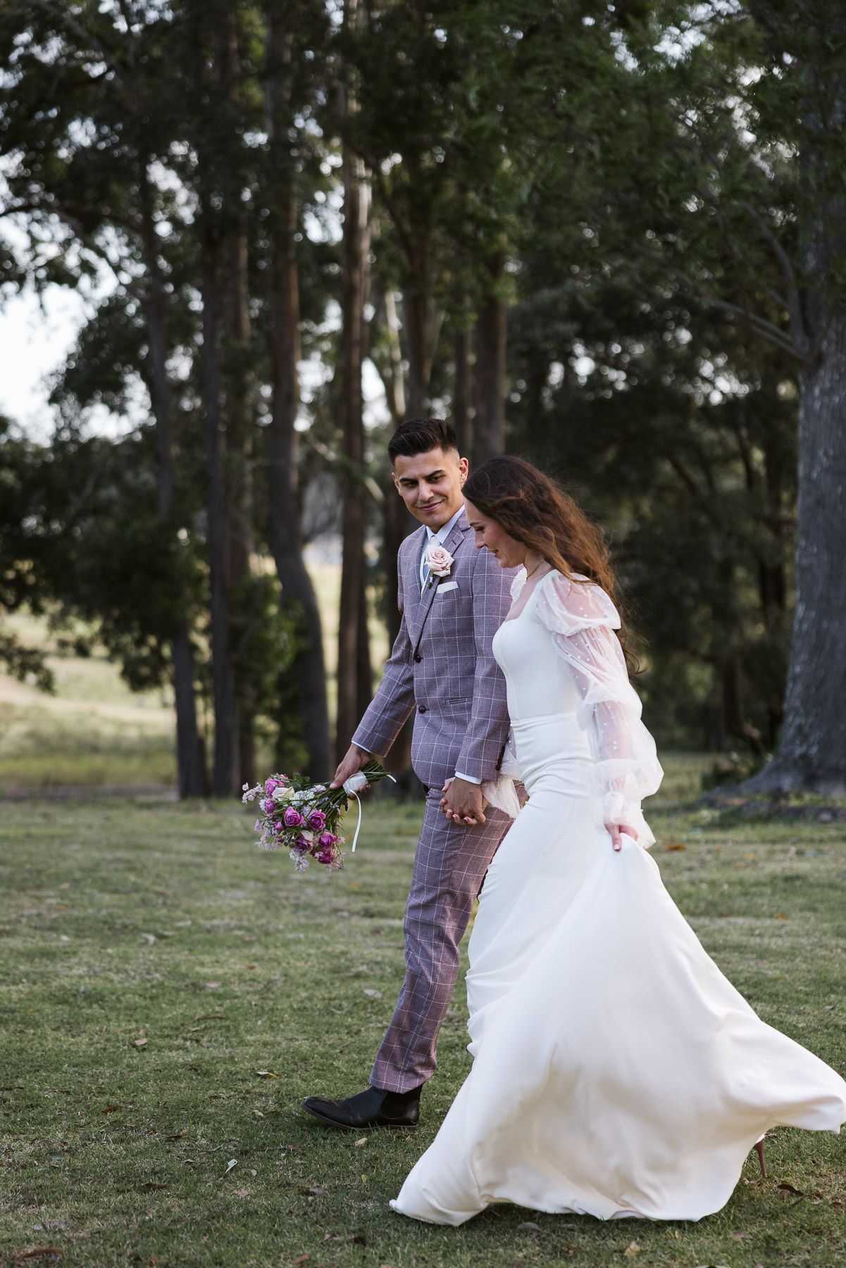 A bride and groom walk hand in hand outdoors, she in a flowing white gown, he in a gray suit holding a bouquet.