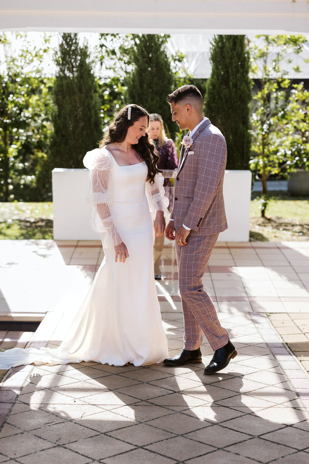 Bride in white gown and groom in plaid suit holding hands at outdoor wedding ceremony with greenery backdrop.