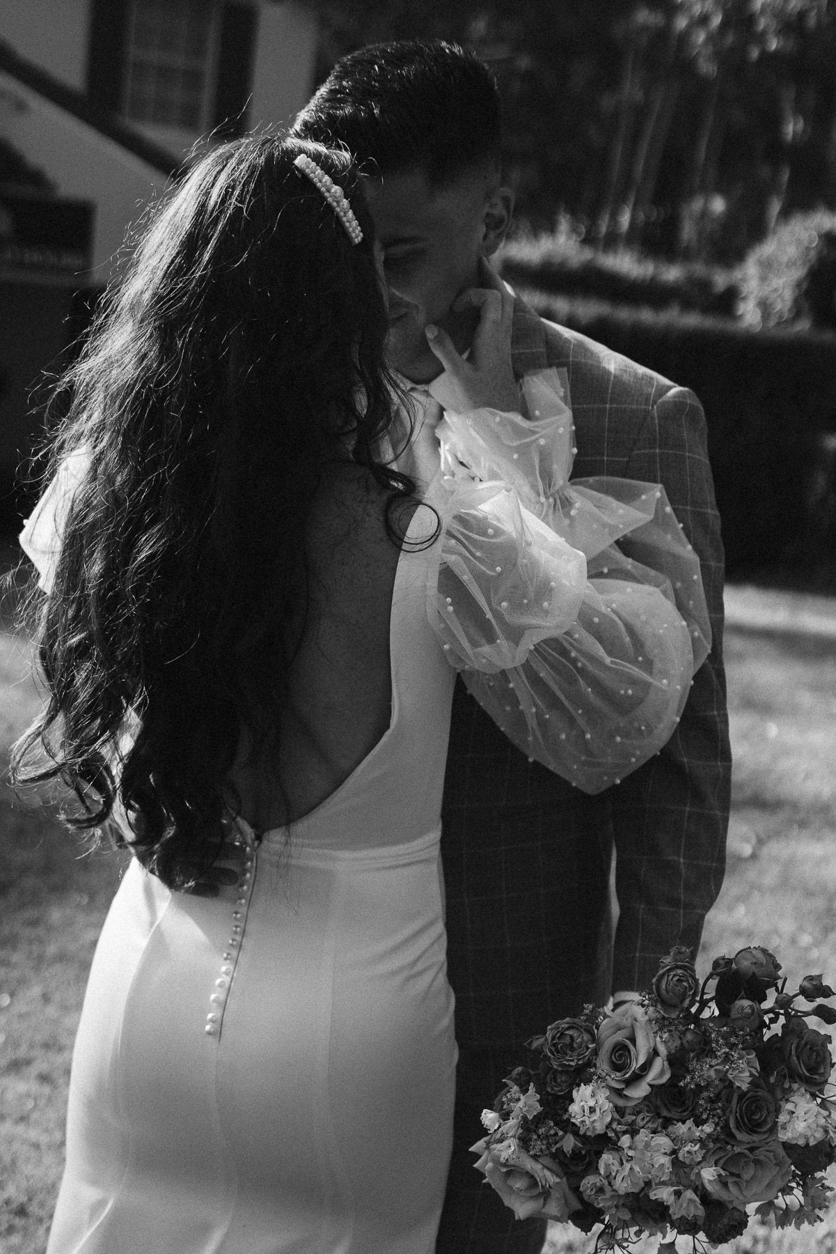 Black and white photo of bride and groom sharing intimate moment, groom kissing bride holding floral bouquet.