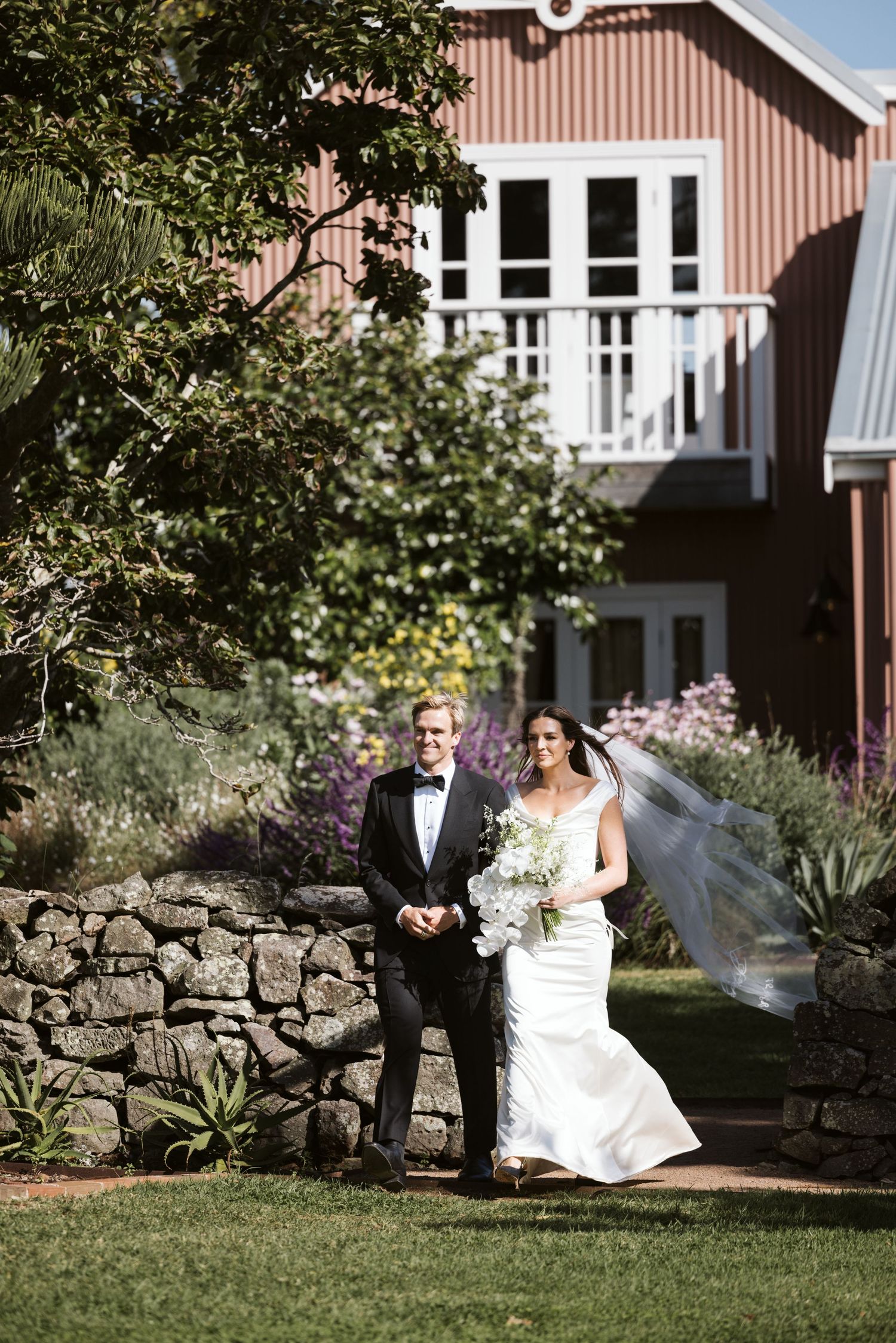 A wedding couple poses in front of a rustic barn-style building with white trim and stone wall landscaping.