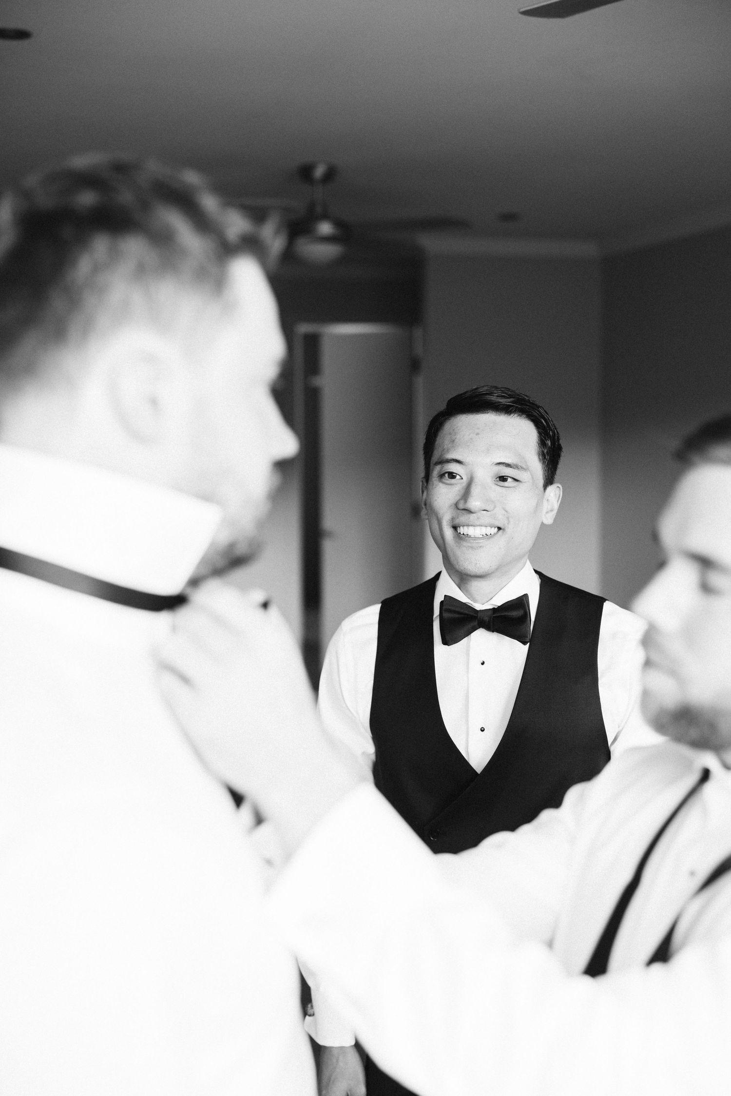 A groomsman in a black vest and bowtie smiles during wedding preparations.
