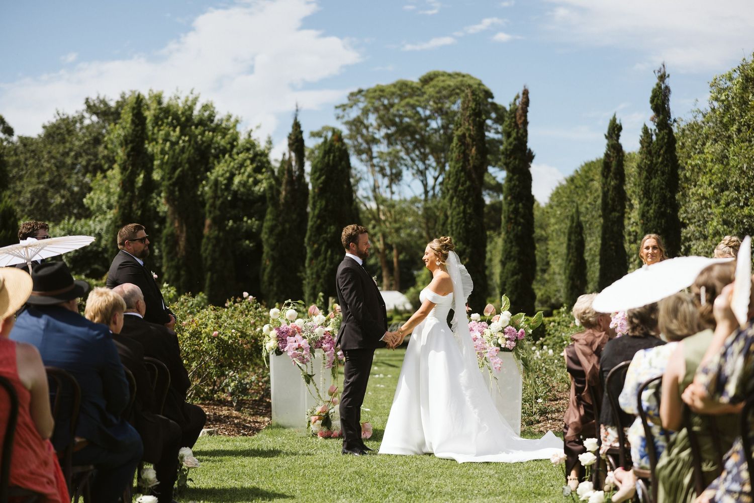 Garden wedding ceremony with guests watching couple exchange vows under cypress trees.