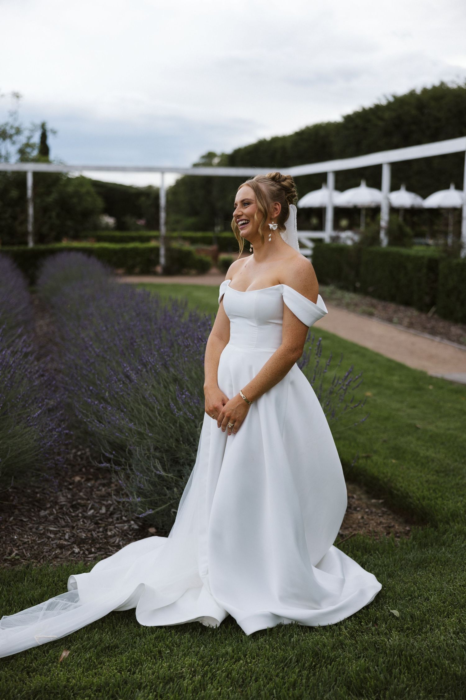 Elegant bride in off-shoulder white gown poses in garden with lavender and white tent in background.