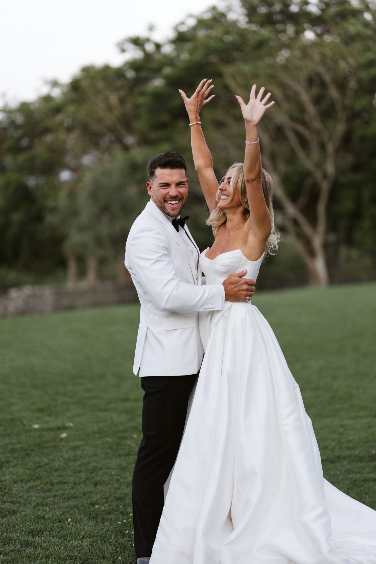 A celebratory moment as a newlywed couple raises their arms in joy on a grassy lawn.