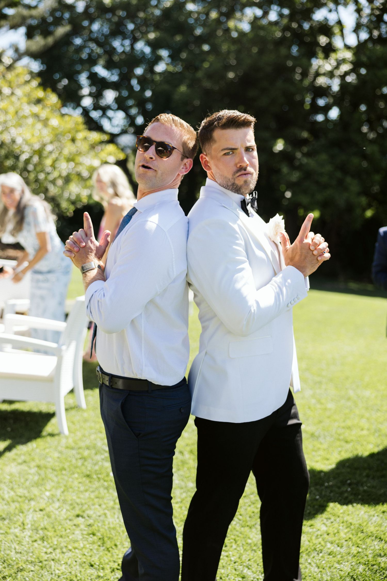 Two groomsmen in white shirts strike a back-to-back pose with peace signs on a sunny lawn.