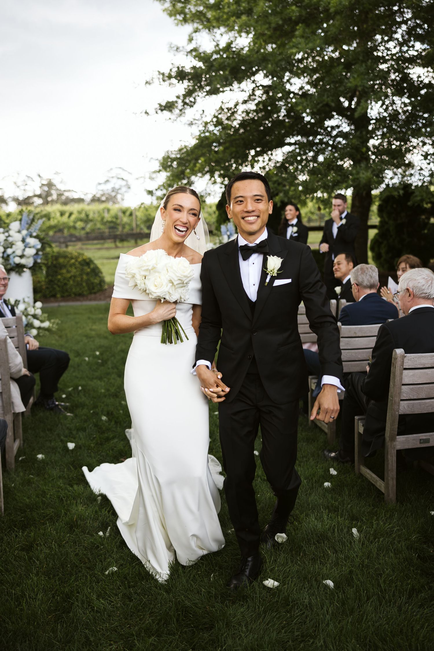 Happy newlyweds walking down the aisle amid guests, bride in fitted white dress holding white bouquet.