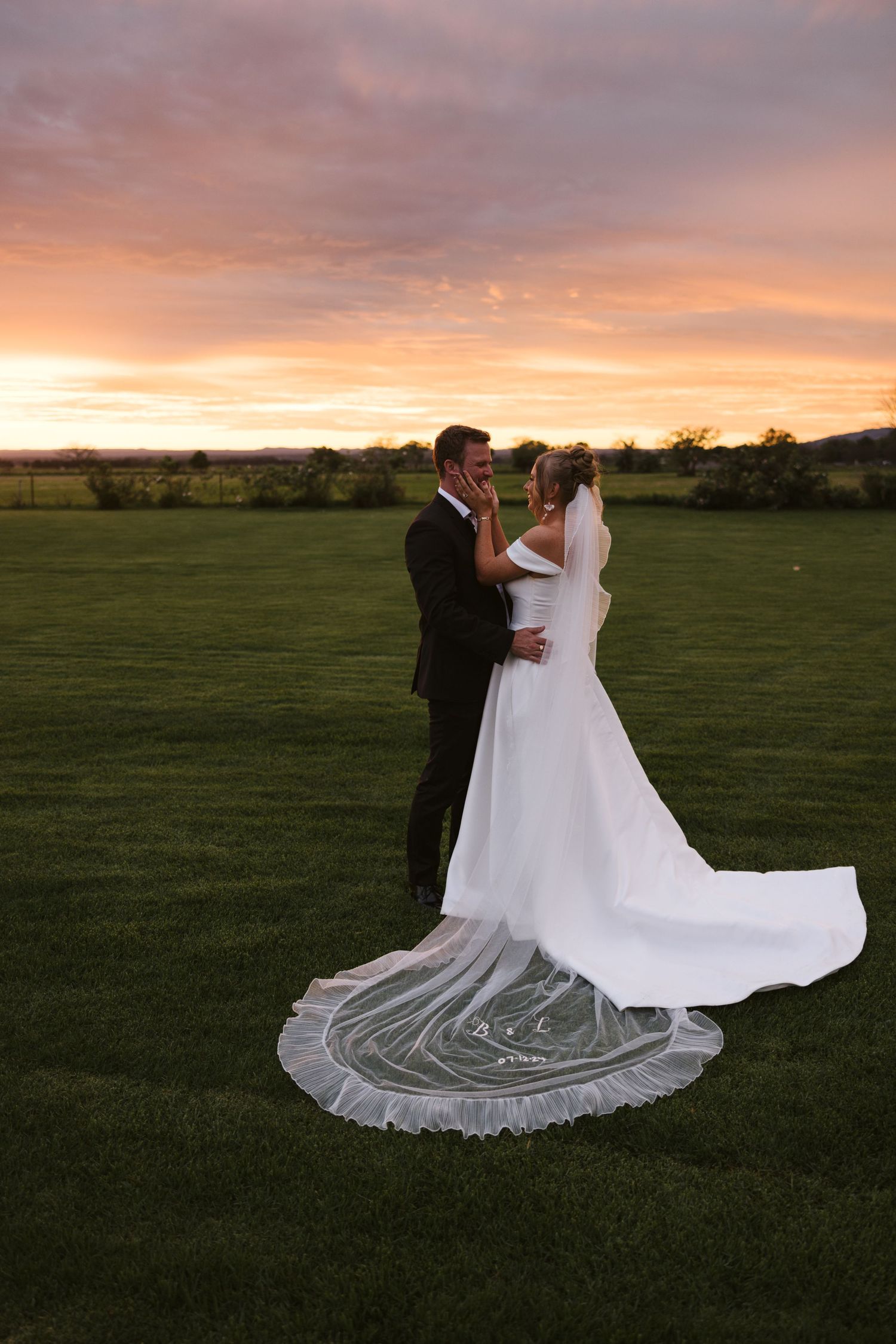 A wedding couple embracing on a grassy field during golden hour sunset.
