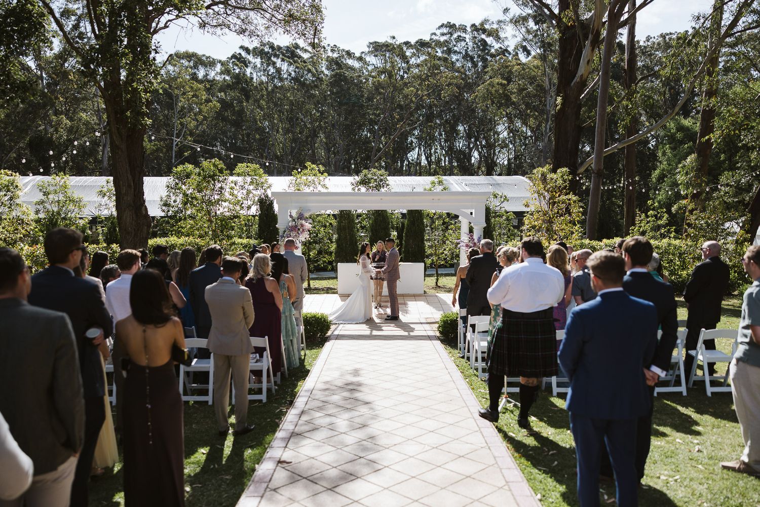 Outdoor wedding ceremony with guests seated on white chairs along a garden aisle leading to a floral arbor.
