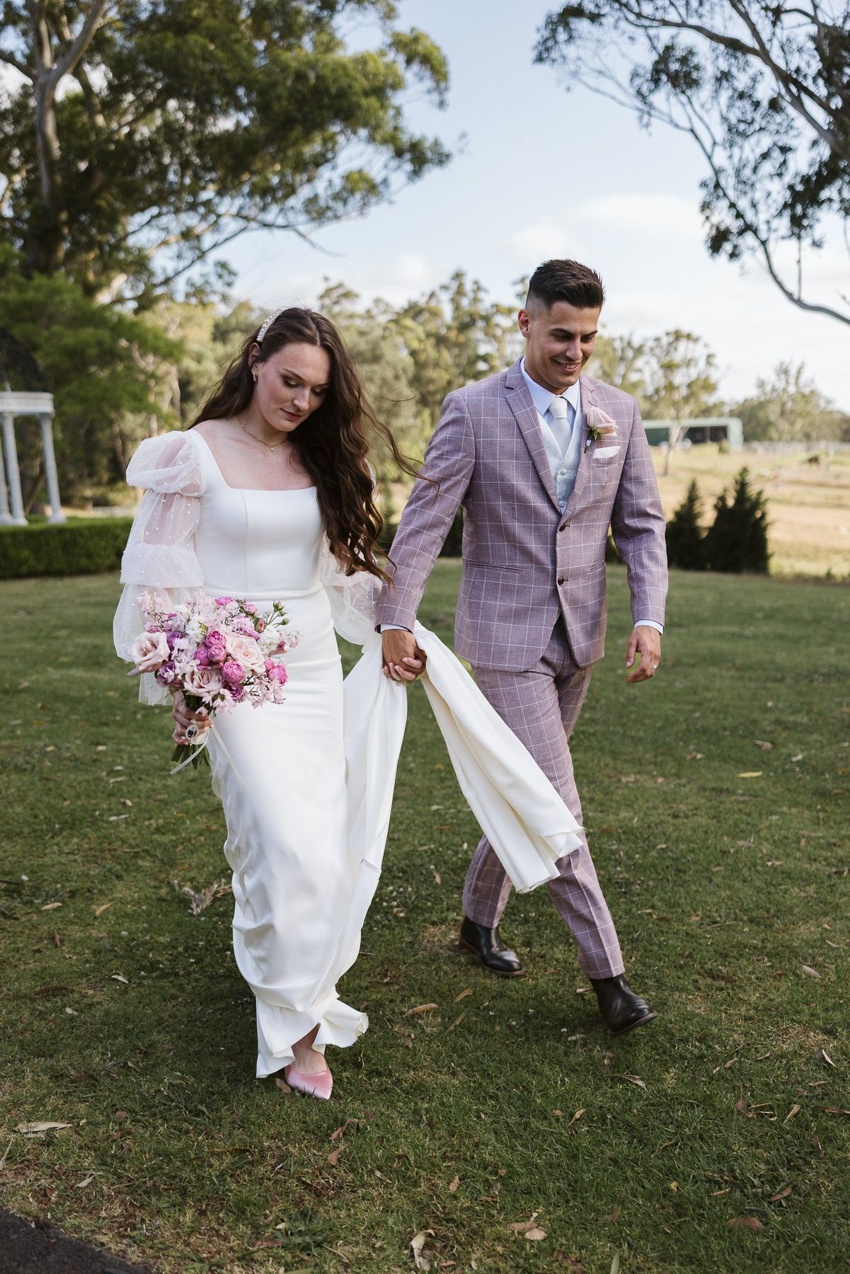 Bride in white gown holding pink bouquet walks with groom in pink plaid suit on green lawn at outdoor wedding.