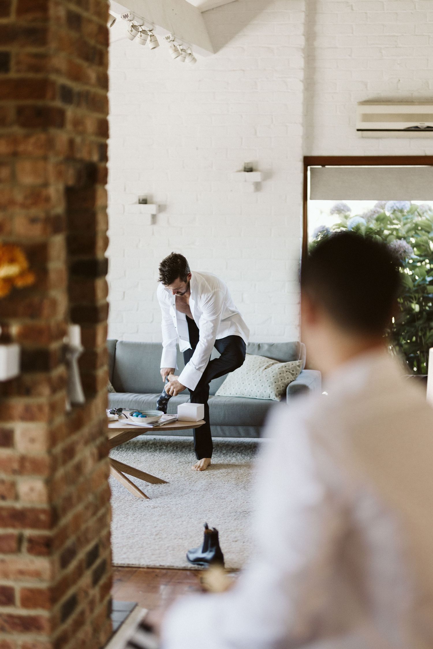 A person in white dress shirt puts on formal shoes while seated on a gray couch in a modern room.