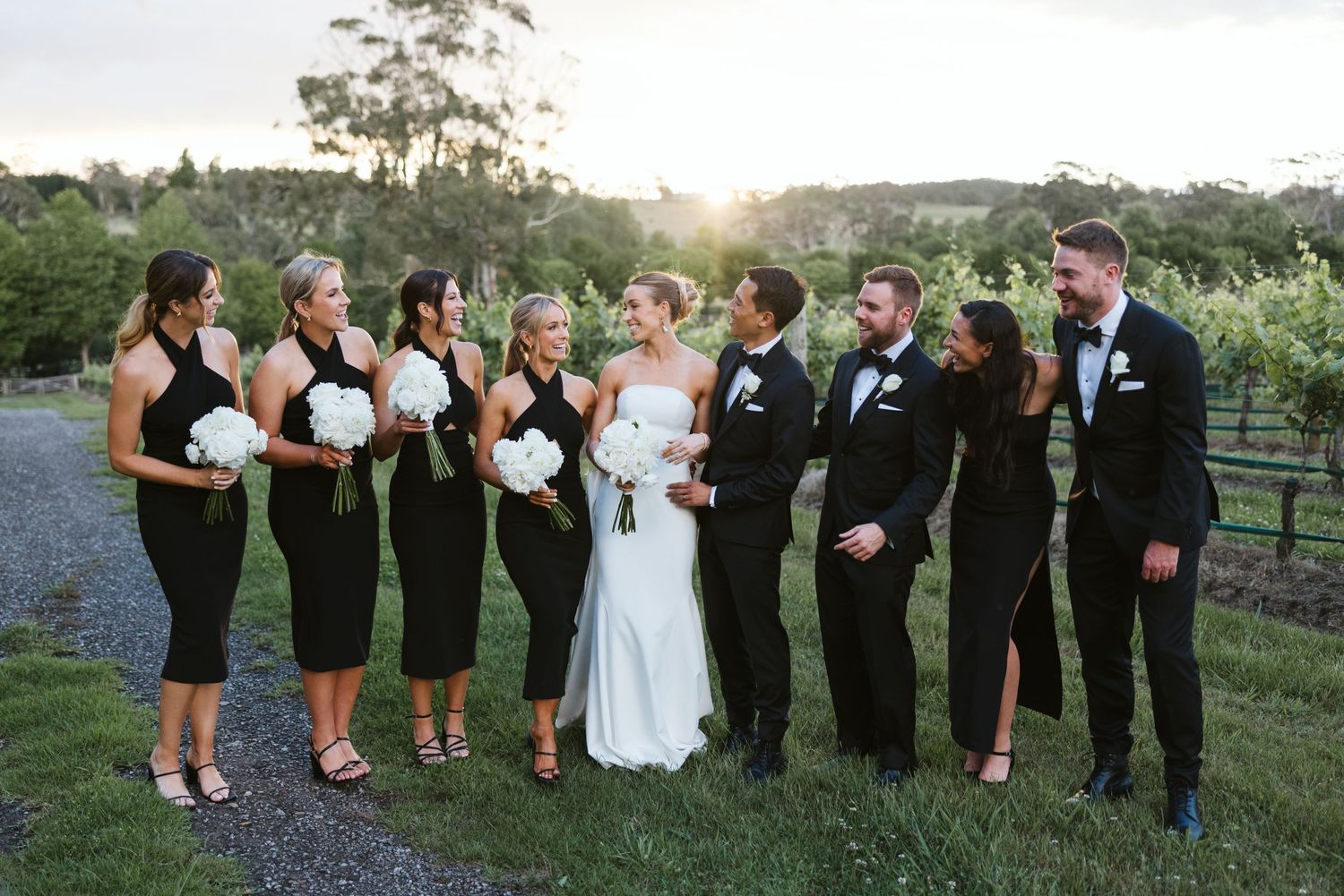 Wedding party in black attire poses together on gravel path near vineyard.