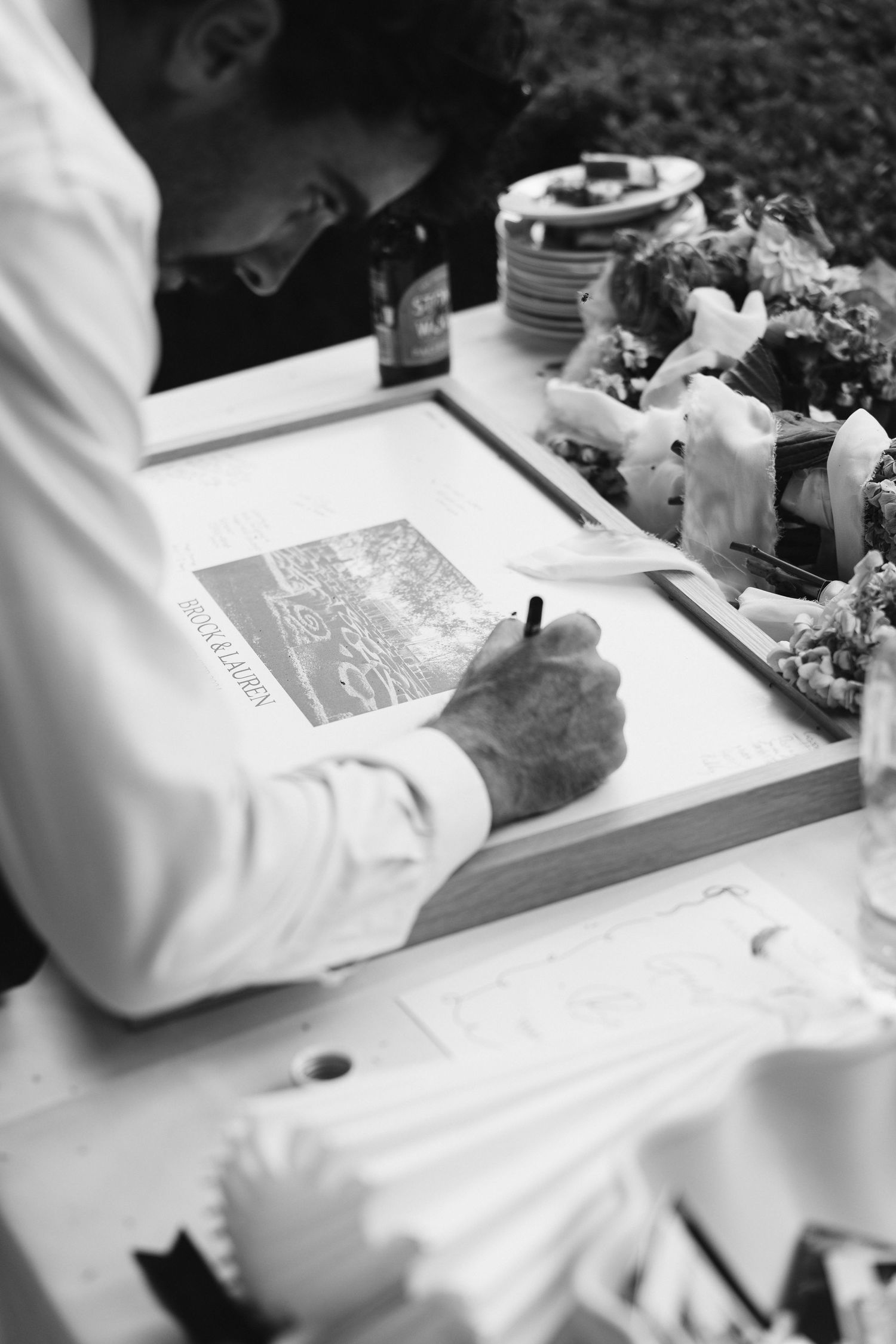 Black and white image of a person signing a guest book at a table with decorative items.
