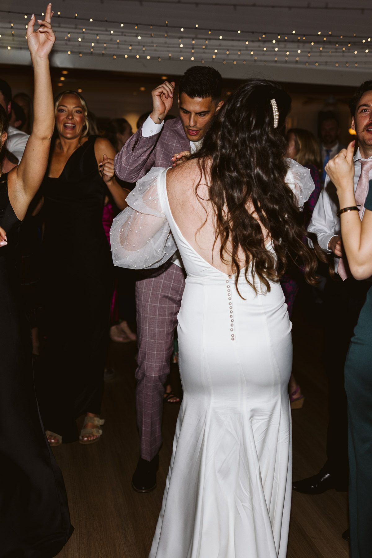 Bride in white gown dancing with groom in plaid suit at wedding reception with guests celebrating.