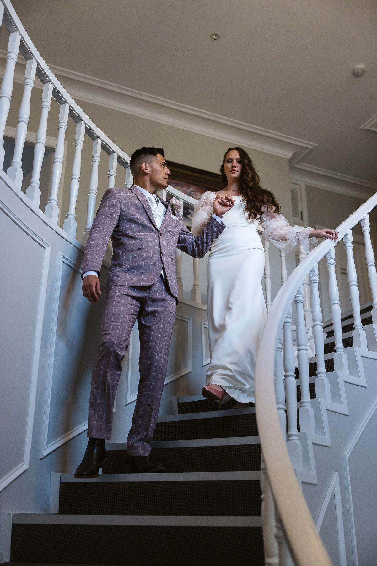 Bride and groom descend elegant white staircase, she in white gown, he in plaid suit, holding hands.