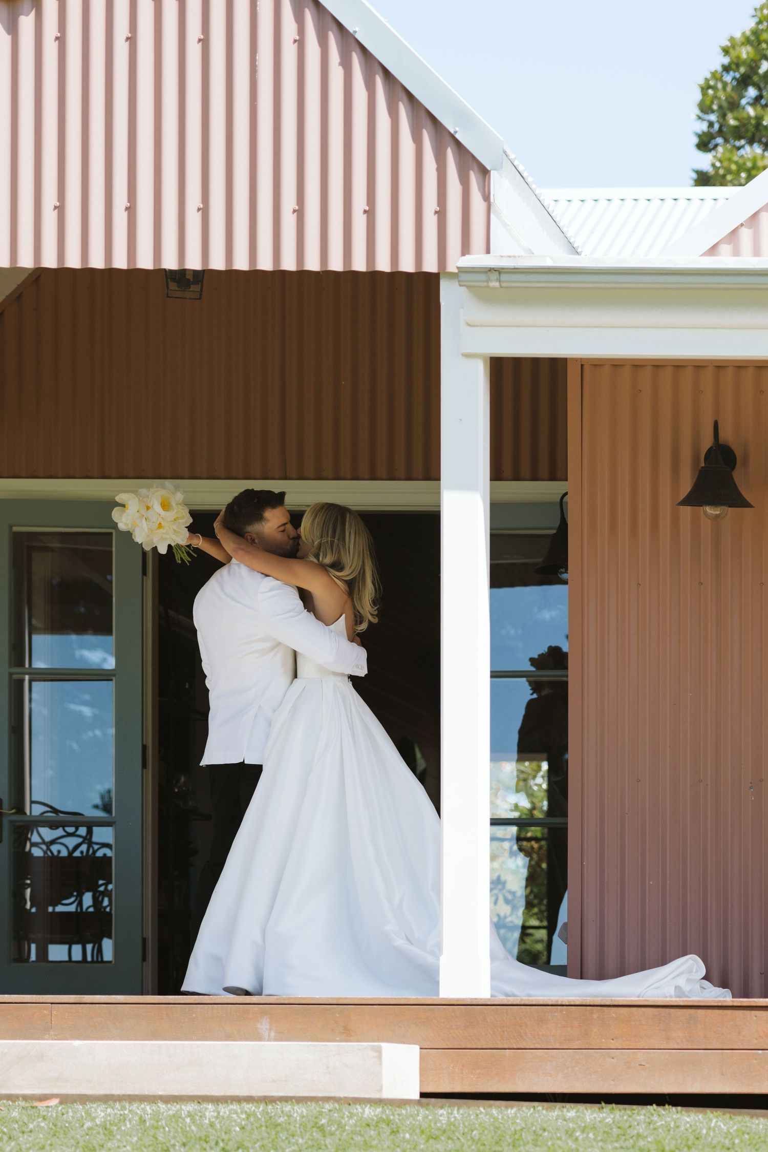 Newlyweds share an intimate moment on a rustic porch of a farmhouse-style building.