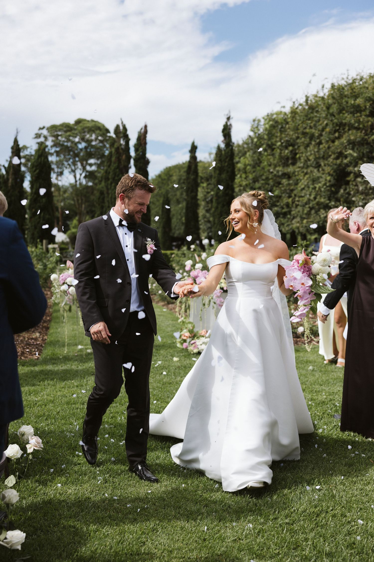 Wedding couple celebrates amid falling flower petals in beautiful garden setting.
