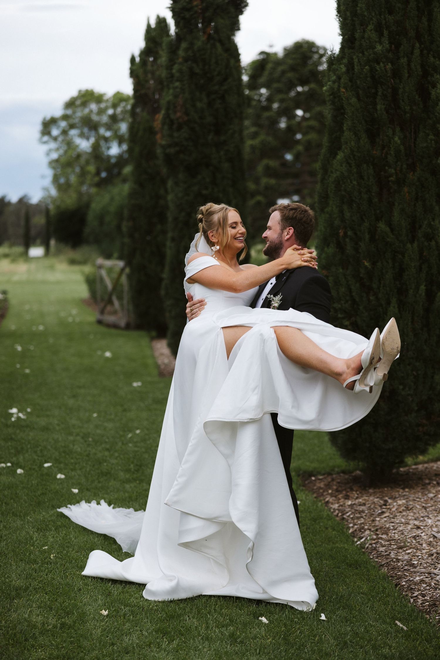 Joyful wedding moment in a garden as groom lifts bride in elegant white dress beside cypress trees.