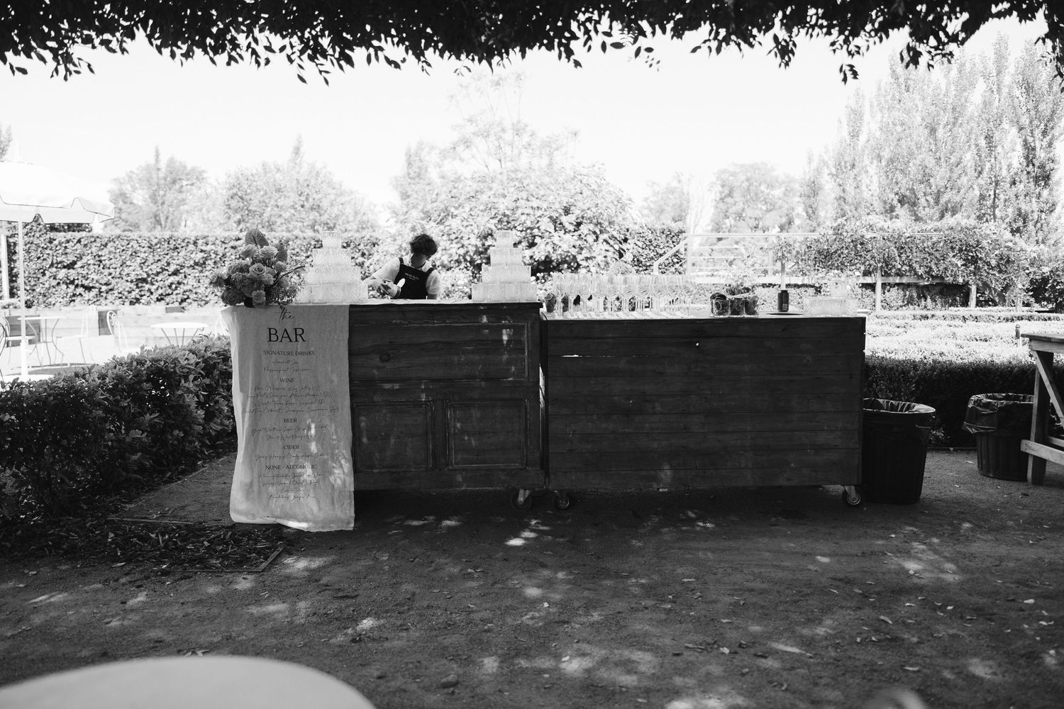 Rustic wooden bar setup with glassware under shaded trees at an outdoor event.