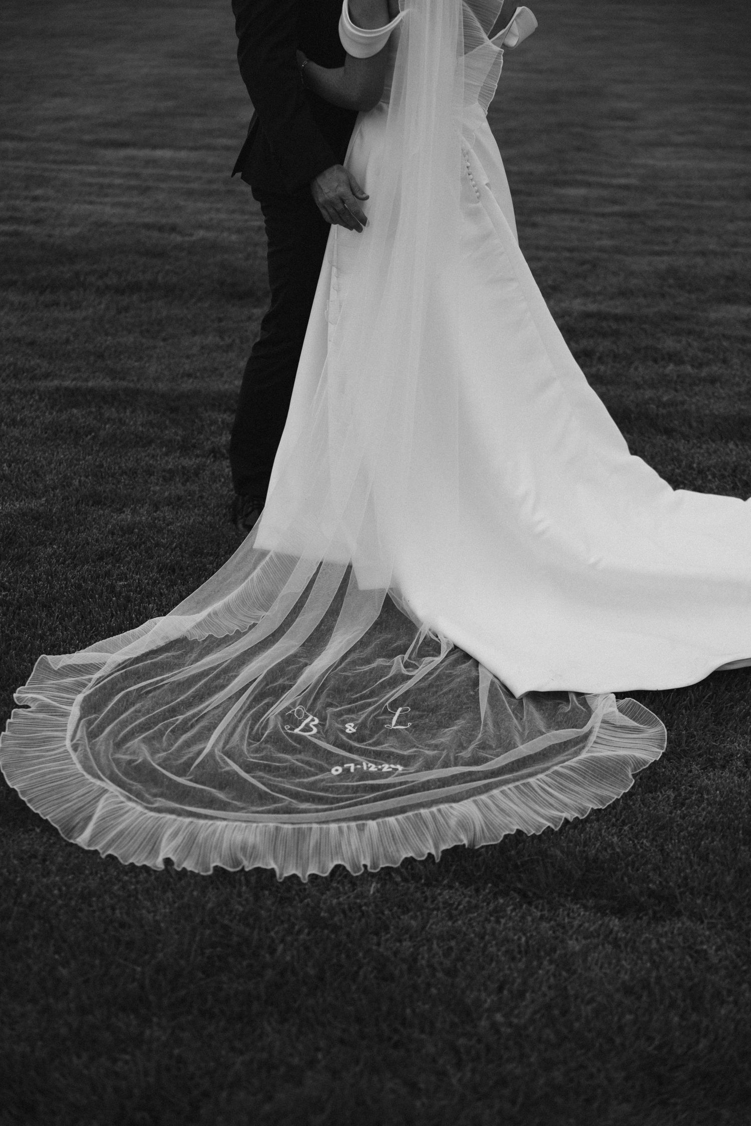 A black and white photo of an elegant wedding dress train with lace details laying on grass.