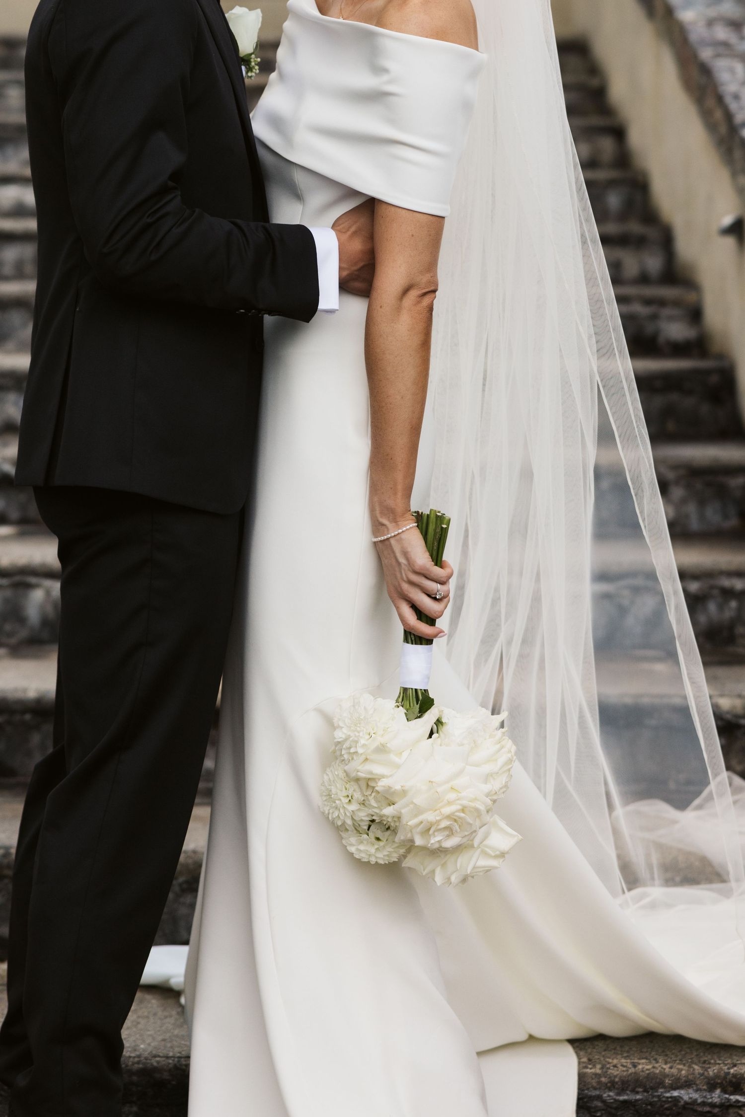 Close-up detail of wedding couple holding white bouquet on stone steps with flowing bridal gown and veil.
