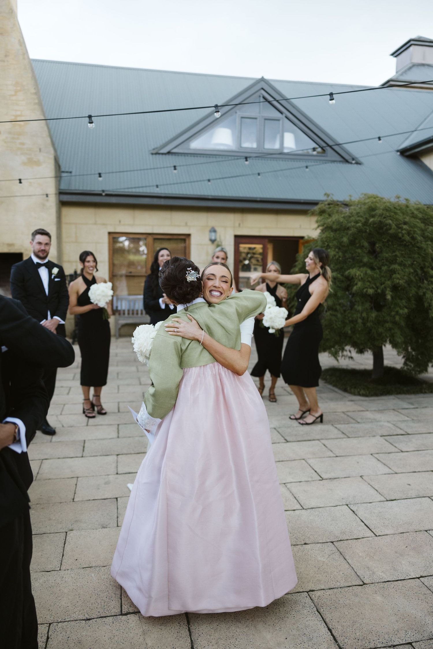 Wedding couple embraces in playful moment on stone patio with string lights and wedding party looking on.