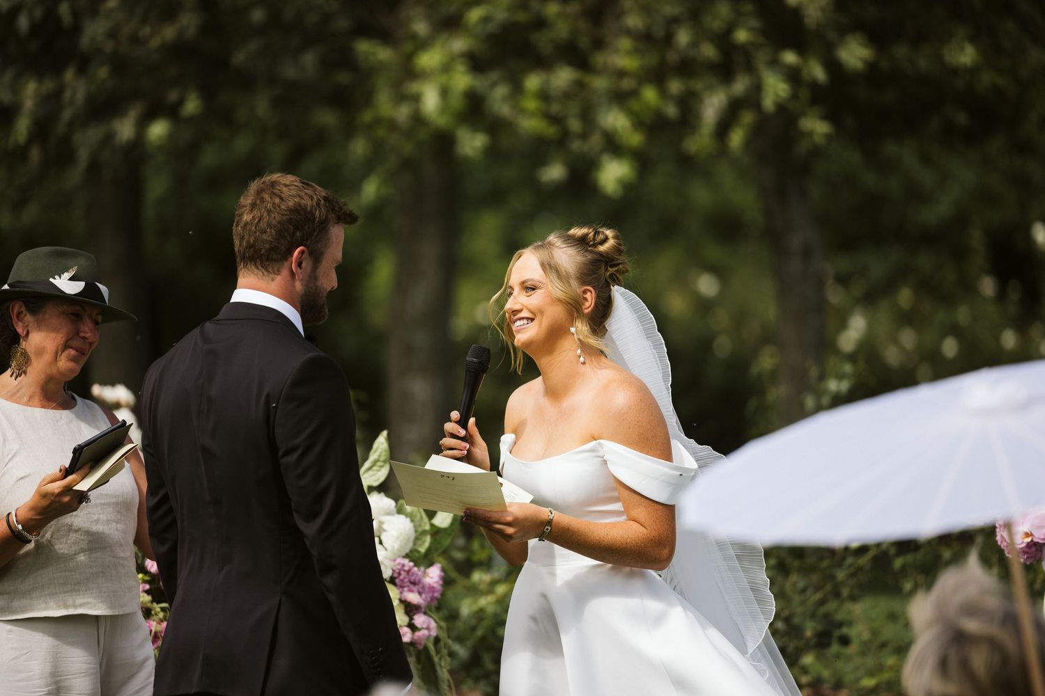 A joyful outdoor wedding ceremony with bride and groom exchanging vows beneath trees.