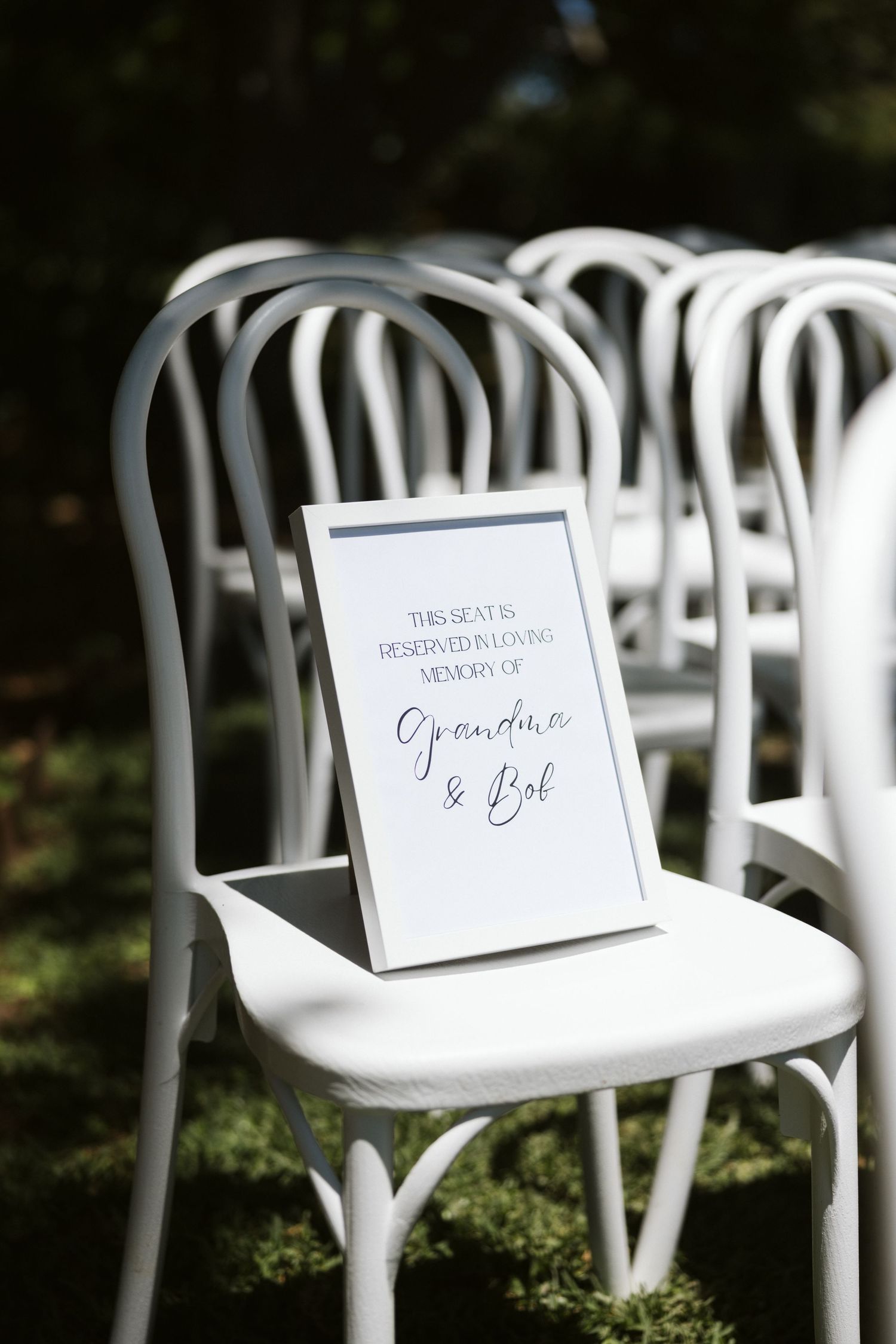 A wedding welcome sign in a black frame rests on a white bentwood chair at an outdoor ceremony.