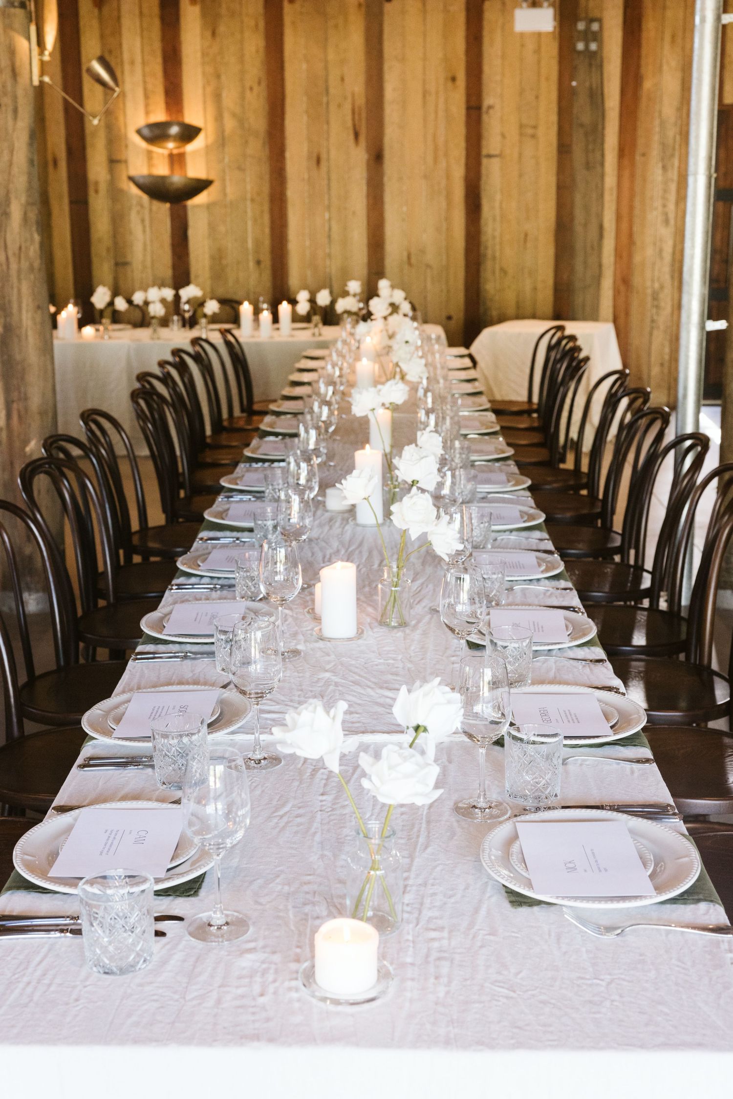 Long banquet table with white linens, black bentwood chairs, and candlelight in a rustic wooden venue.