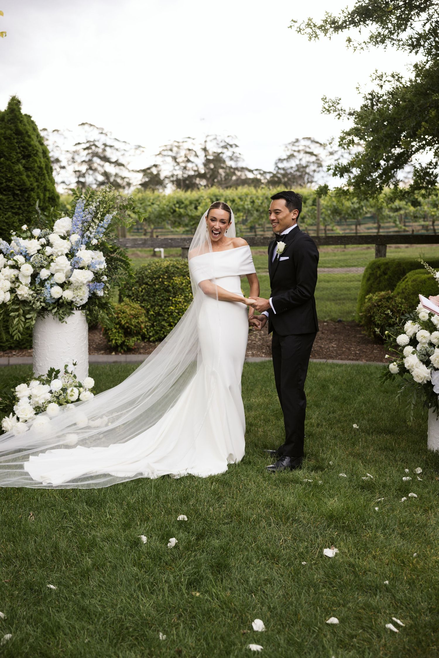 Bride in flowing veil and groom stand by white floral pillars during garden ceremony.