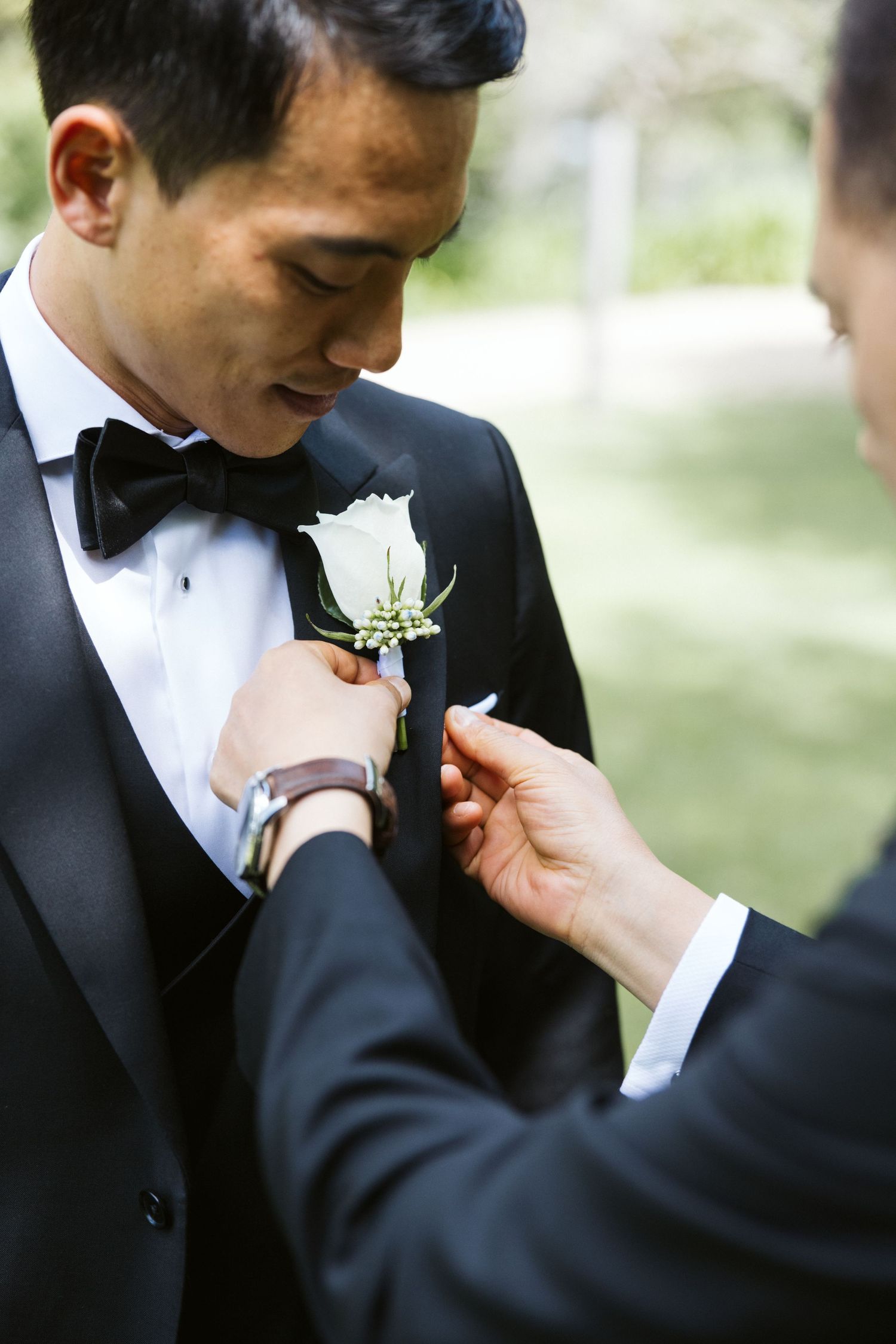 Someone pins a white boutonniere onto a groom's lapel during wedding preparations.