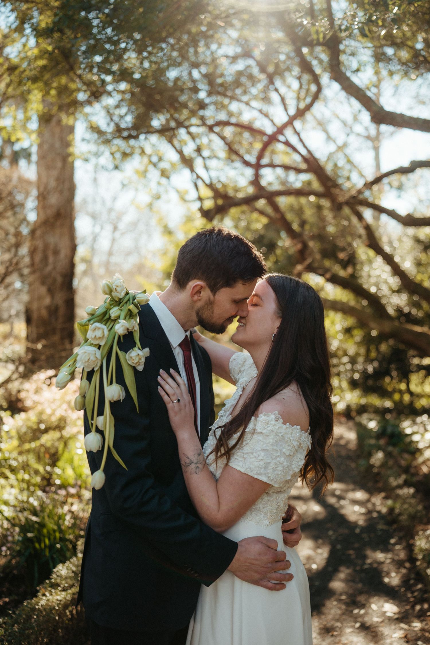 Bride and groom share a romantic kiss surrounded by sunlit trees holding a white tulip bouquet.